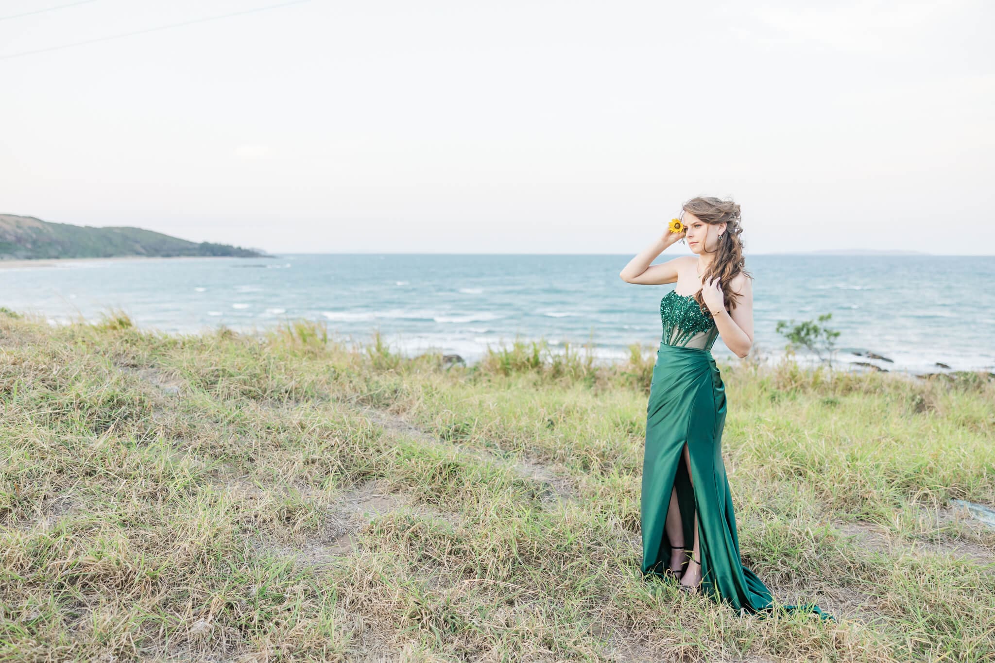 Formal portrait taken near the ocean at Emu Park, Yeppoon, of a graduate girl, in a gorgeous emerald green dress, holding a sunflower to her ear.