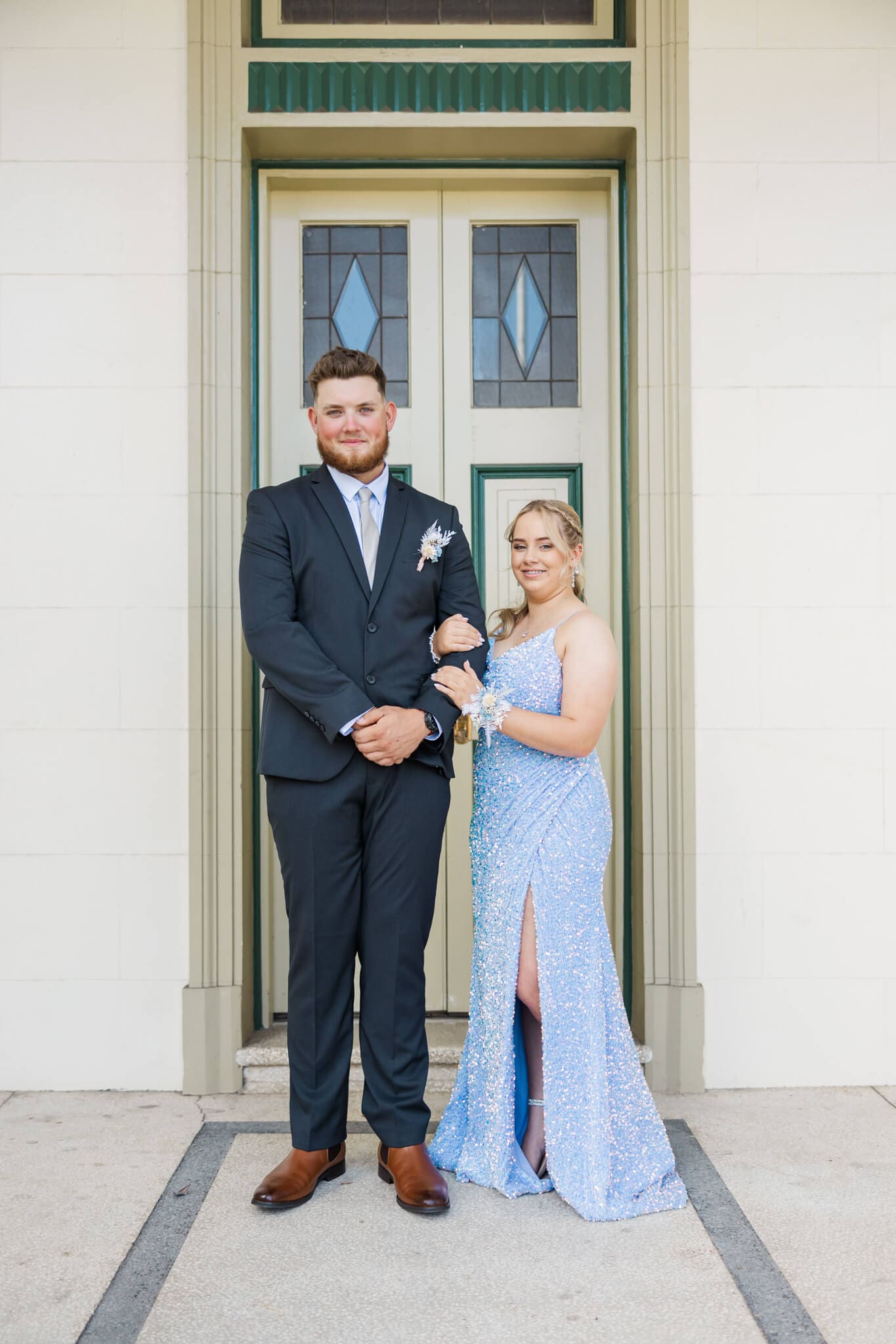 Couple posing in formal dress for graduation photos in a cream building. Girl in sky blue dress holding partner's arm.