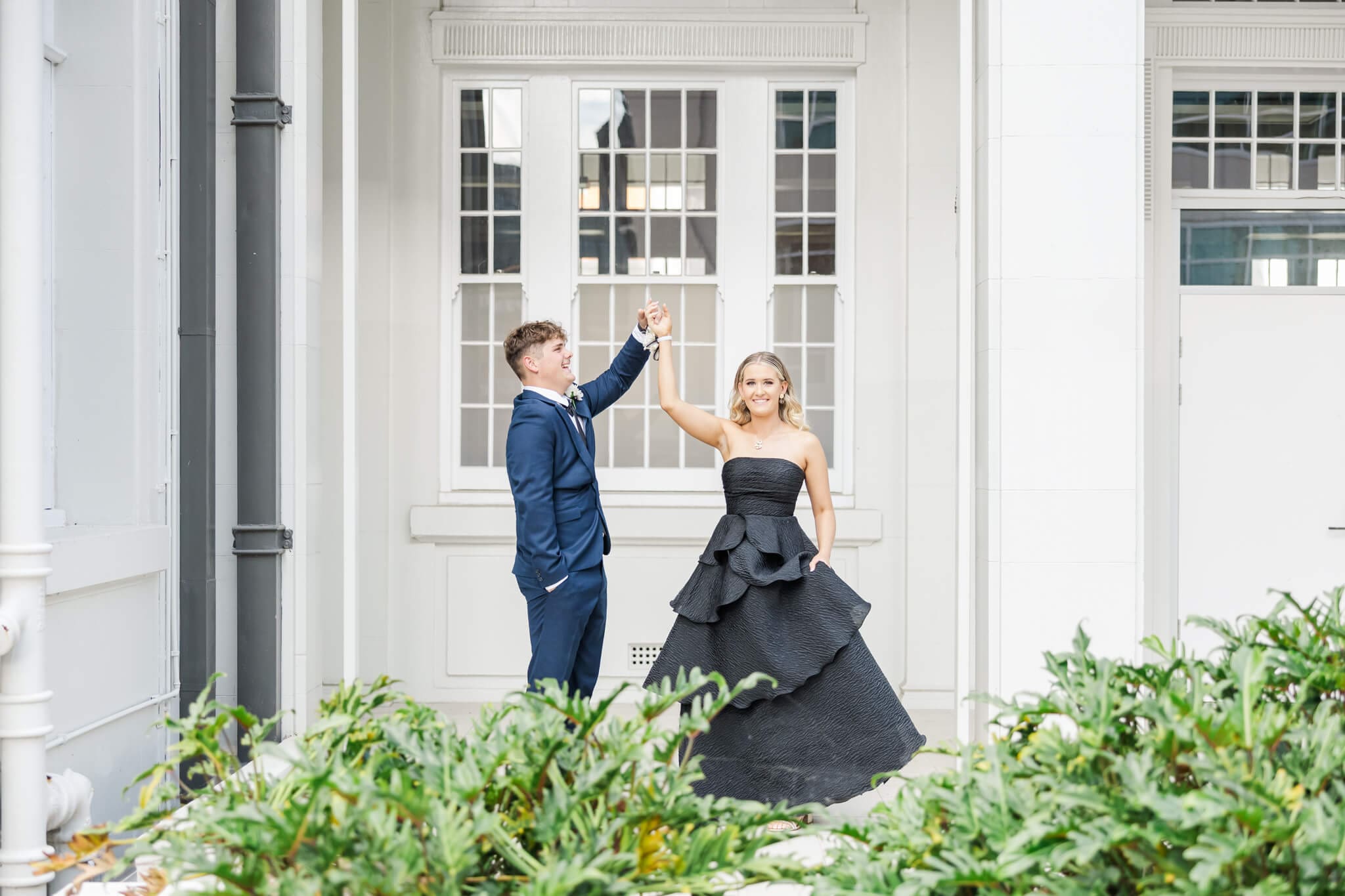 Graduate couple on formal day, dressed in a blue suit and a black, Smoke and Mirrors Boutique dress. Boy spins girl around while Julie-Anne Photography captures the moment.