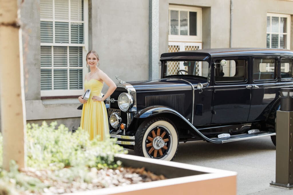 Stunning formal image of girl standing next to an old fashioned black car, dressed in a Gossip Girl dress.