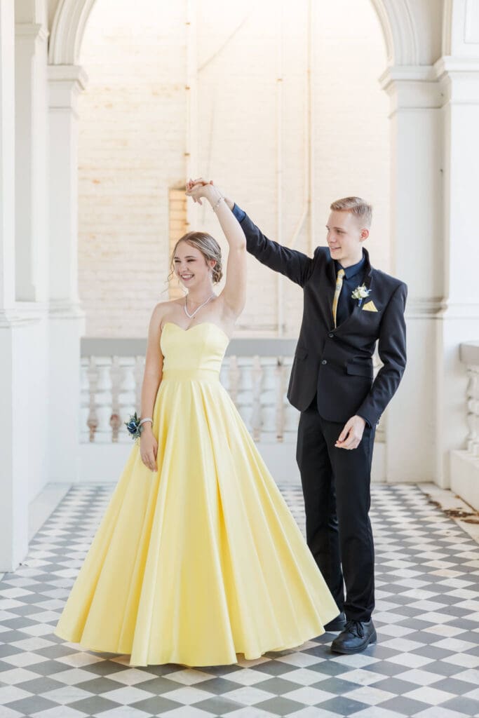Beautiful formal photo of a girl dressed in a yellow formal dress, being twirled around by a boy dressed in a black suit. Photographed by Julie-Anne Photography.