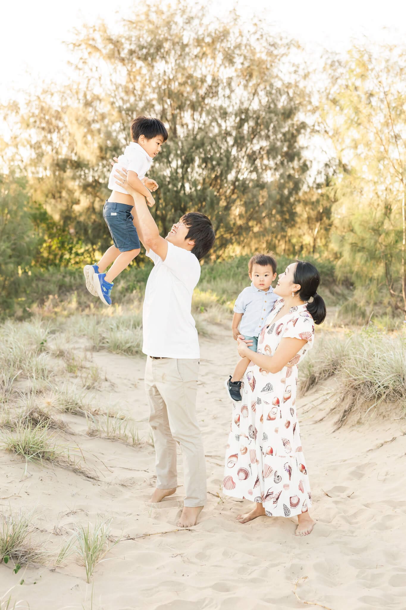 A portrait during a family photo session of Dad throwing his little boy in the air while mum watches on behind while she holds the younger brother.
