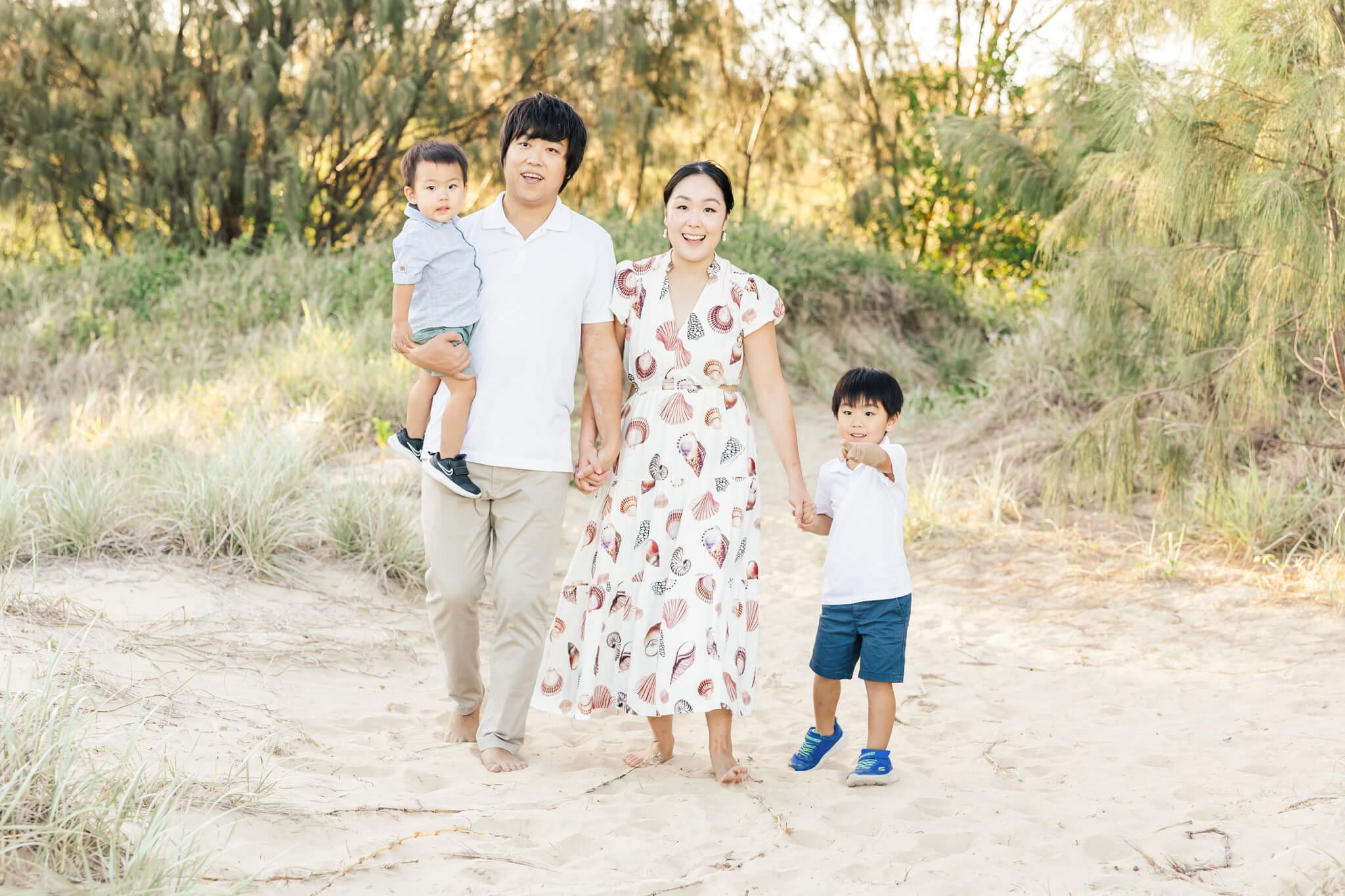 A beautiful beach family photo session capturing a family of 4 - Dad, Mum and 2 little boys, walking in the sand with the beach grass and trees in the background.