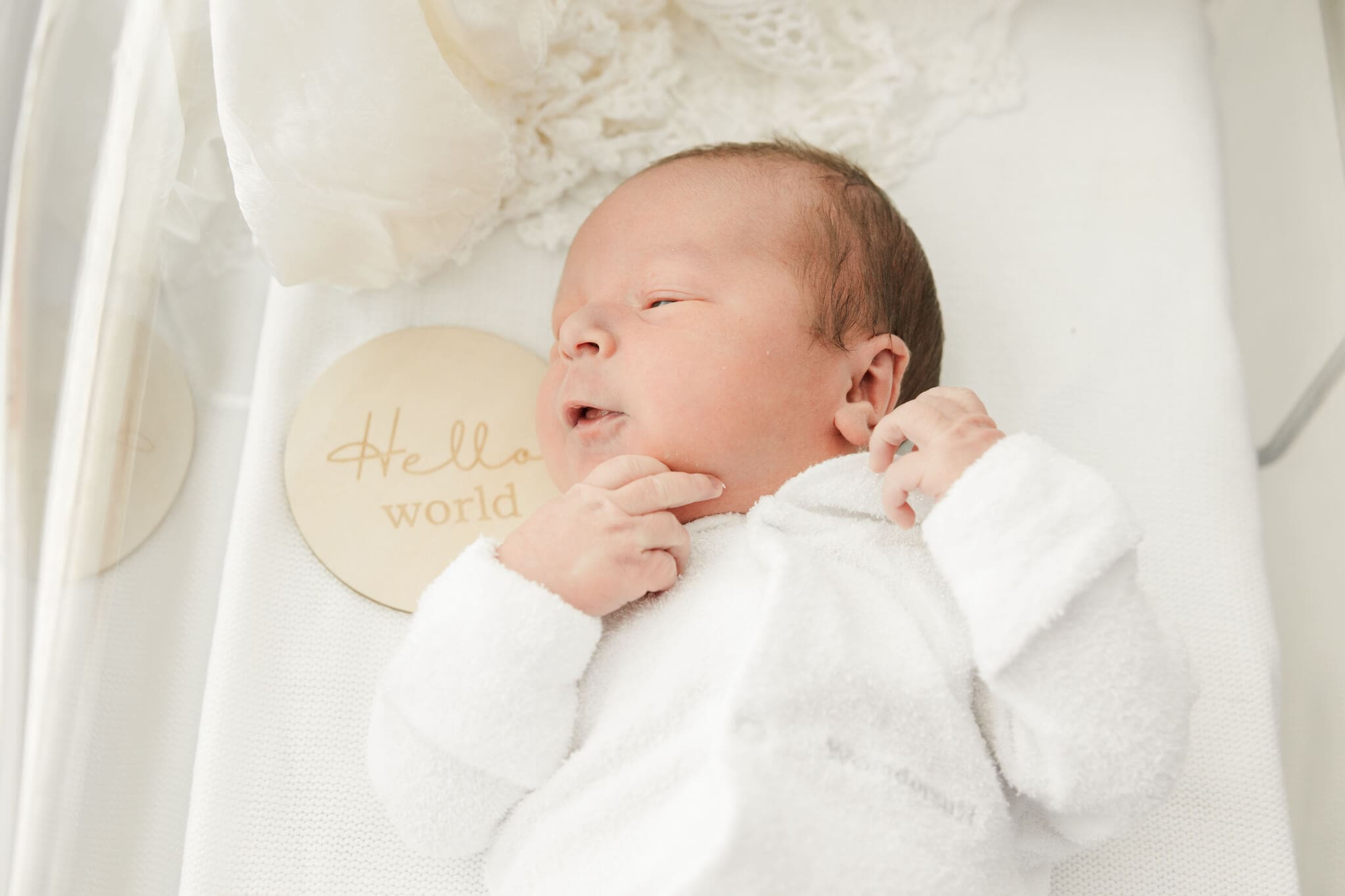 Newborn baby in a crib, lying next to wooden sign that says Hello World.