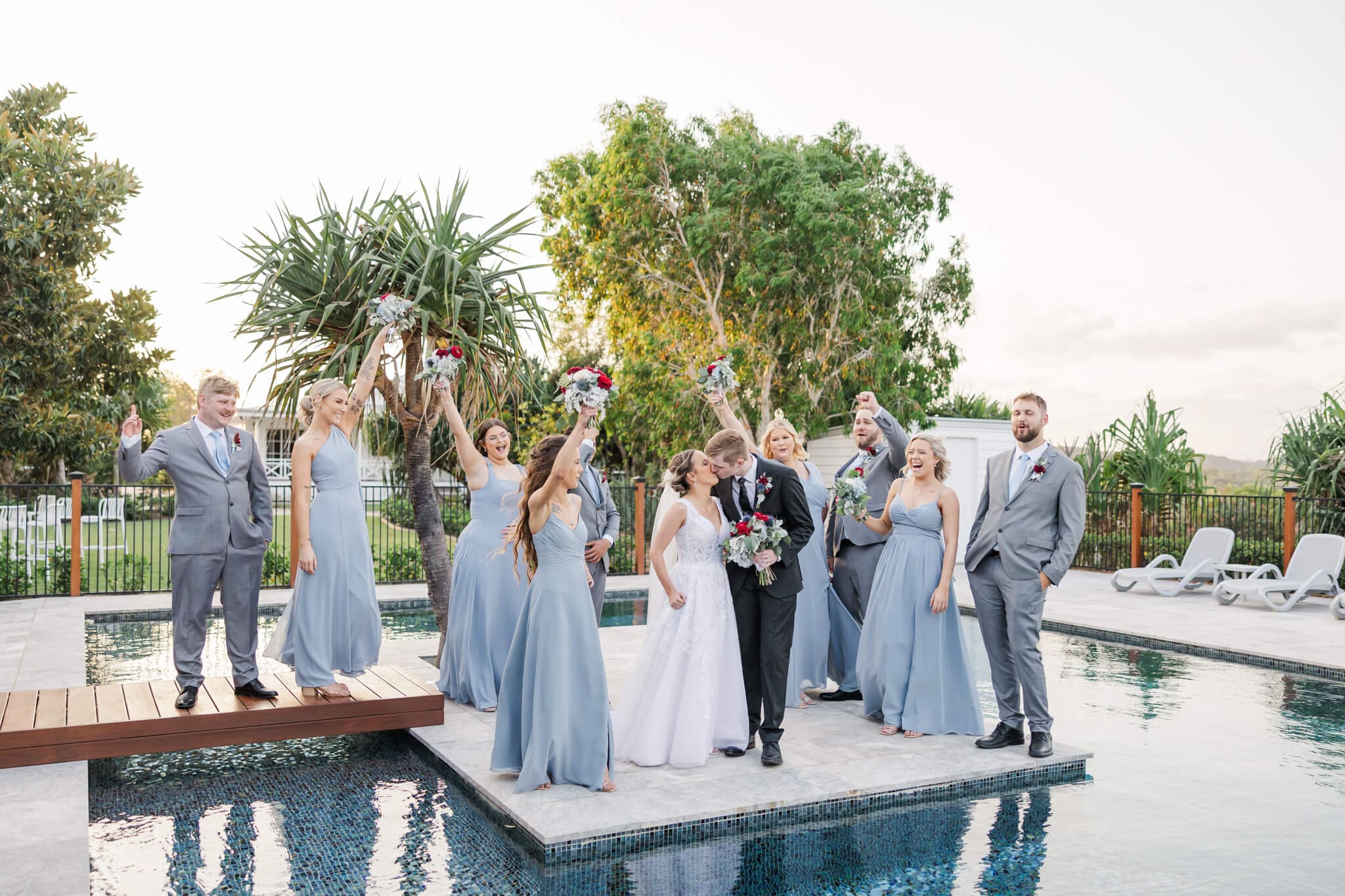 Wedding party celebrate at wedding venue Ambrym Estate Yeppoon as the bride and groom kiss on the pool deck over a pool.