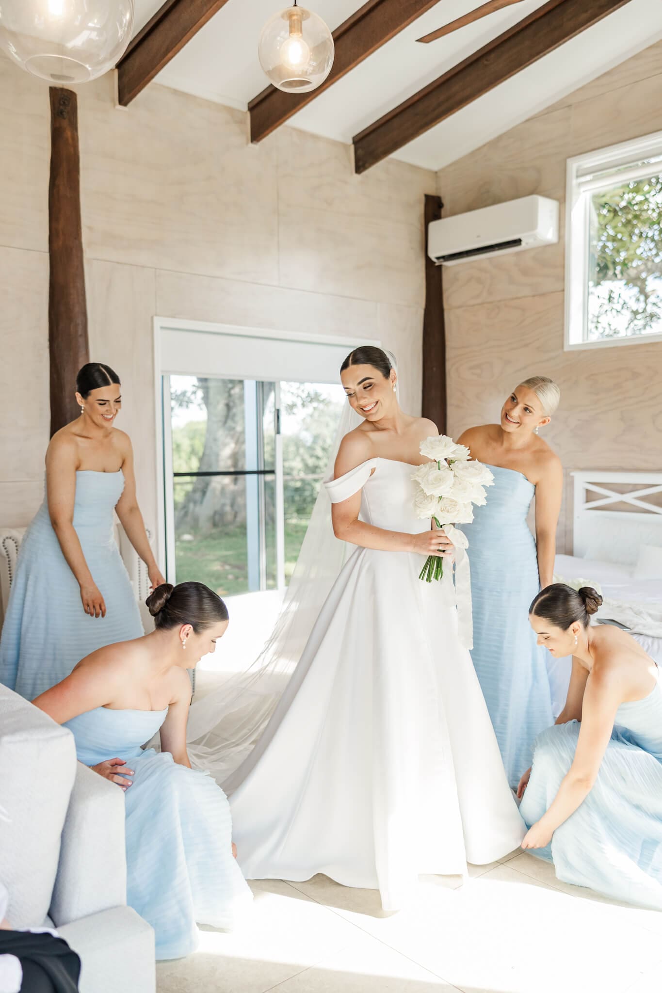A bride glances over her shoulder as the bridesmaids fluff her dress ready for her wedding. Located at The Lookout Woodbury and captured by Julie-Anne Photography.