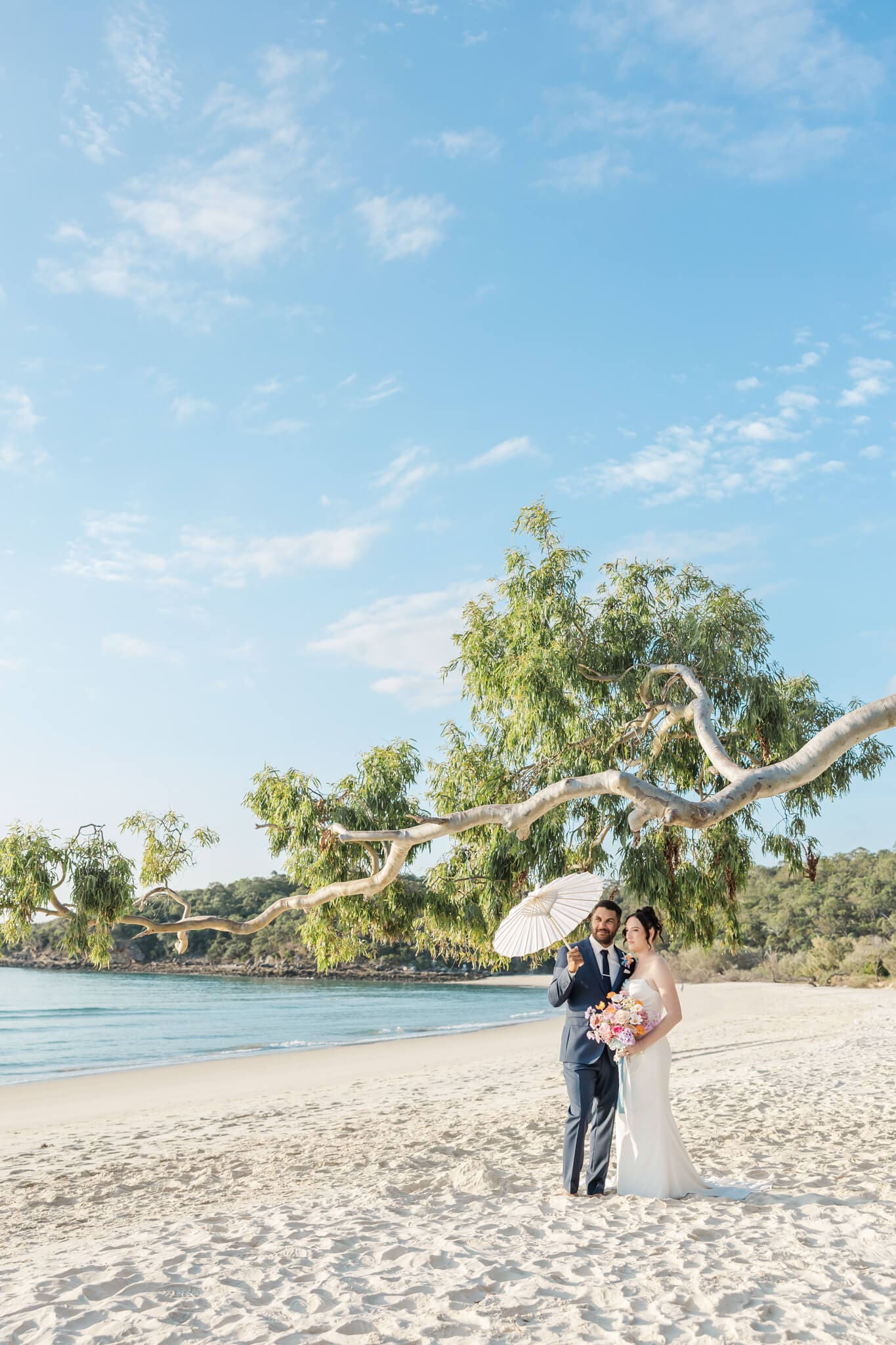 Stunning bride and groom portrait on a beach located at Great Keppel Island Hideaway. Groom holds umbrella over them both while the couple look out over the ocean.