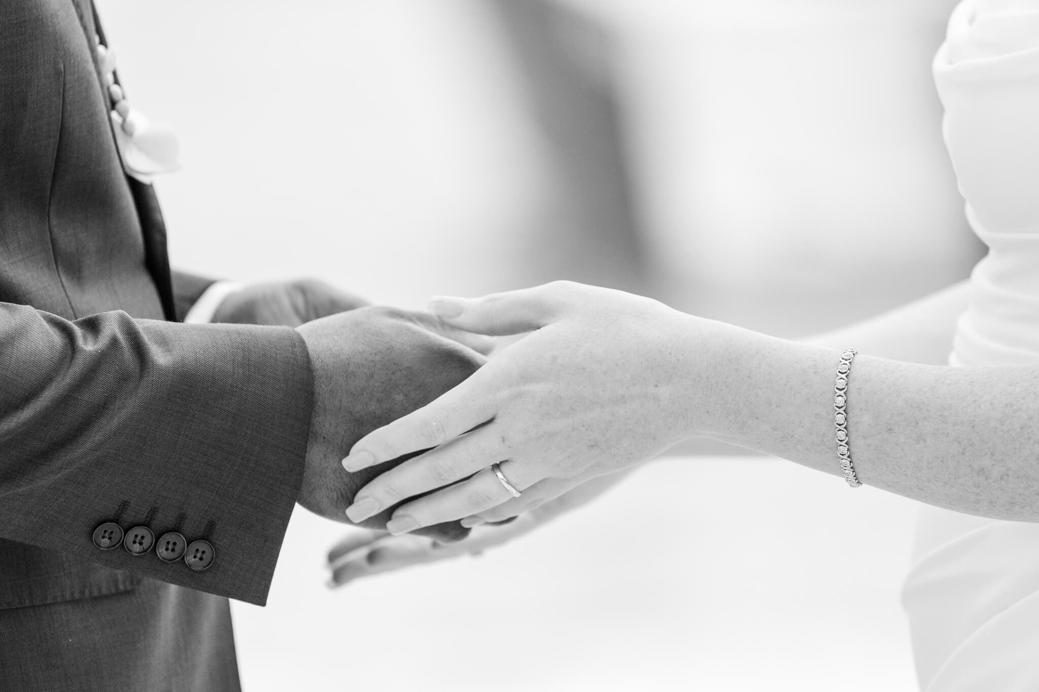 Black and white photo, close up, of bride and groom holding hands while exchanging rings.