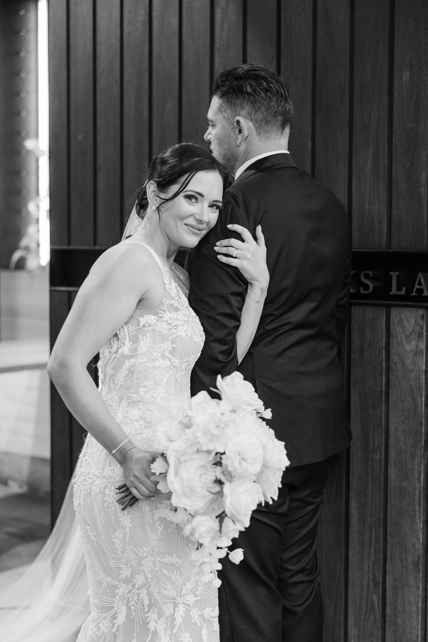 Black and white bridal shot of bride leaning on the groom's shoulder while she smiles at the camera.