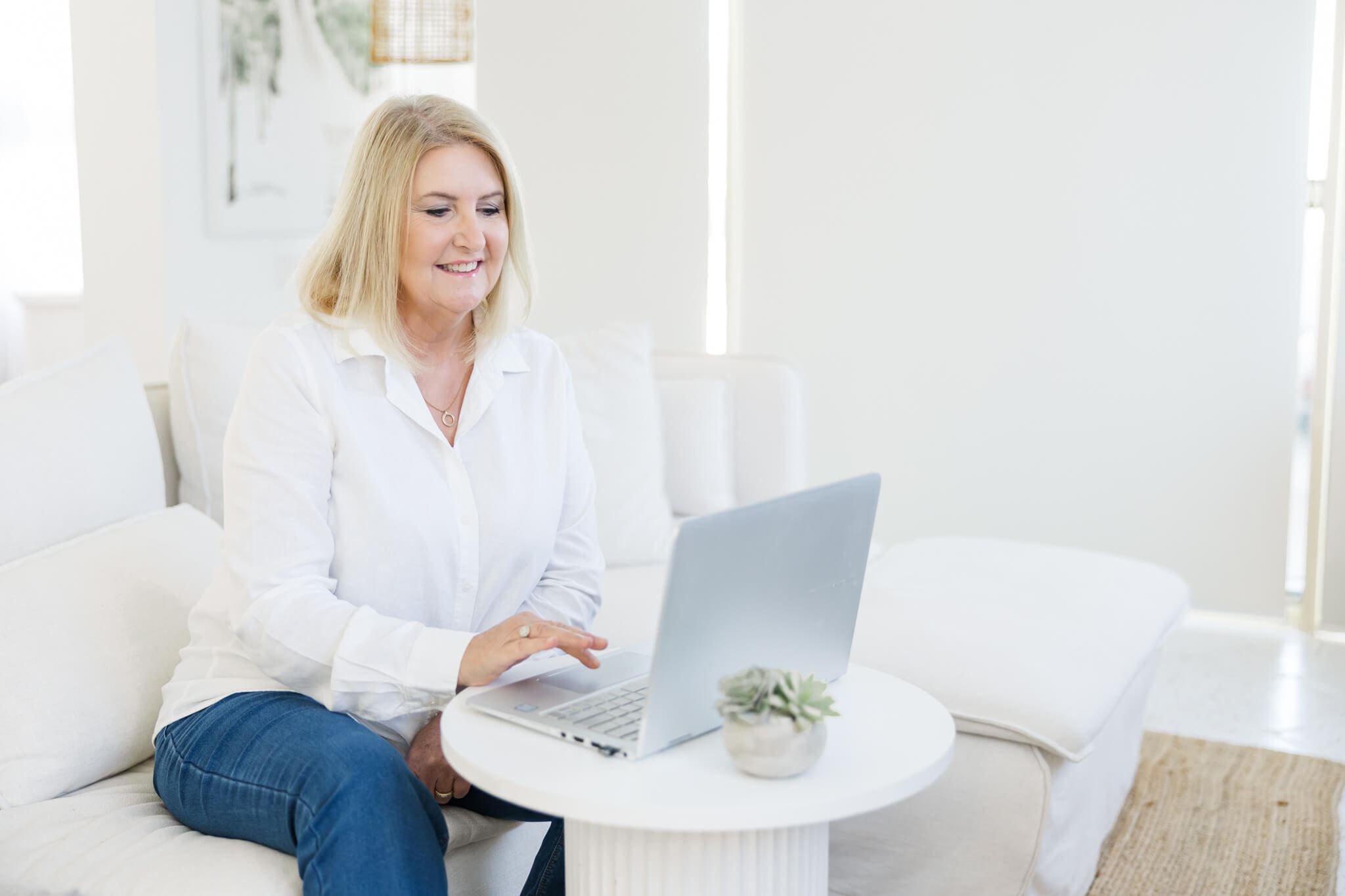 Branding photo of a marriage celebrant dressed in a white blouse and blue jeans, wearing a cream wide brimmed hat, looking at her laptop.