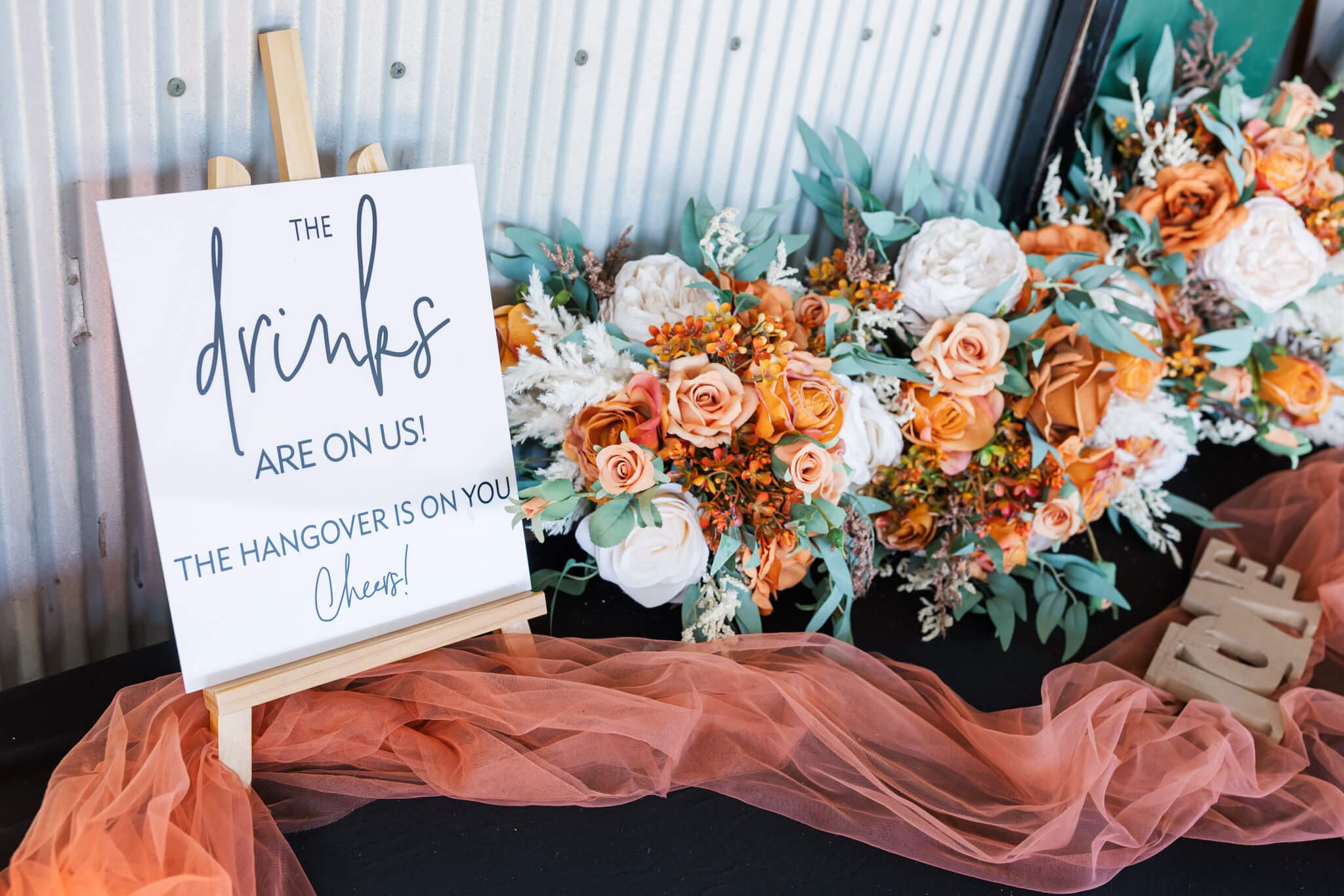 A beautiful arrangement of flowers next to a sign at a wedding that reads 'drinks are on us, the hangover is on you. Cheers!'