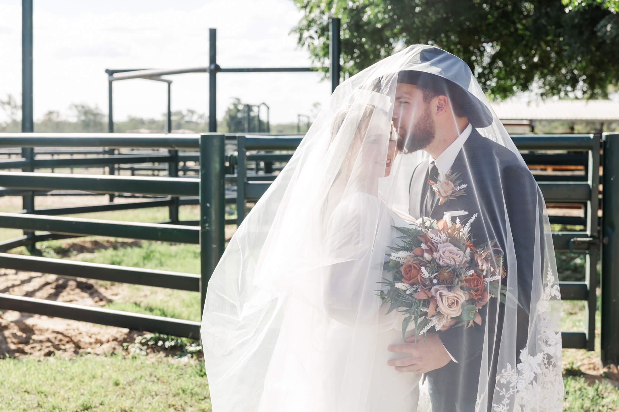 Bride and Groom portrait near the yards at Paradise Lagoons Rockhampton. Bride has veil draped over both her and her grooms head, the groom is kissing her.