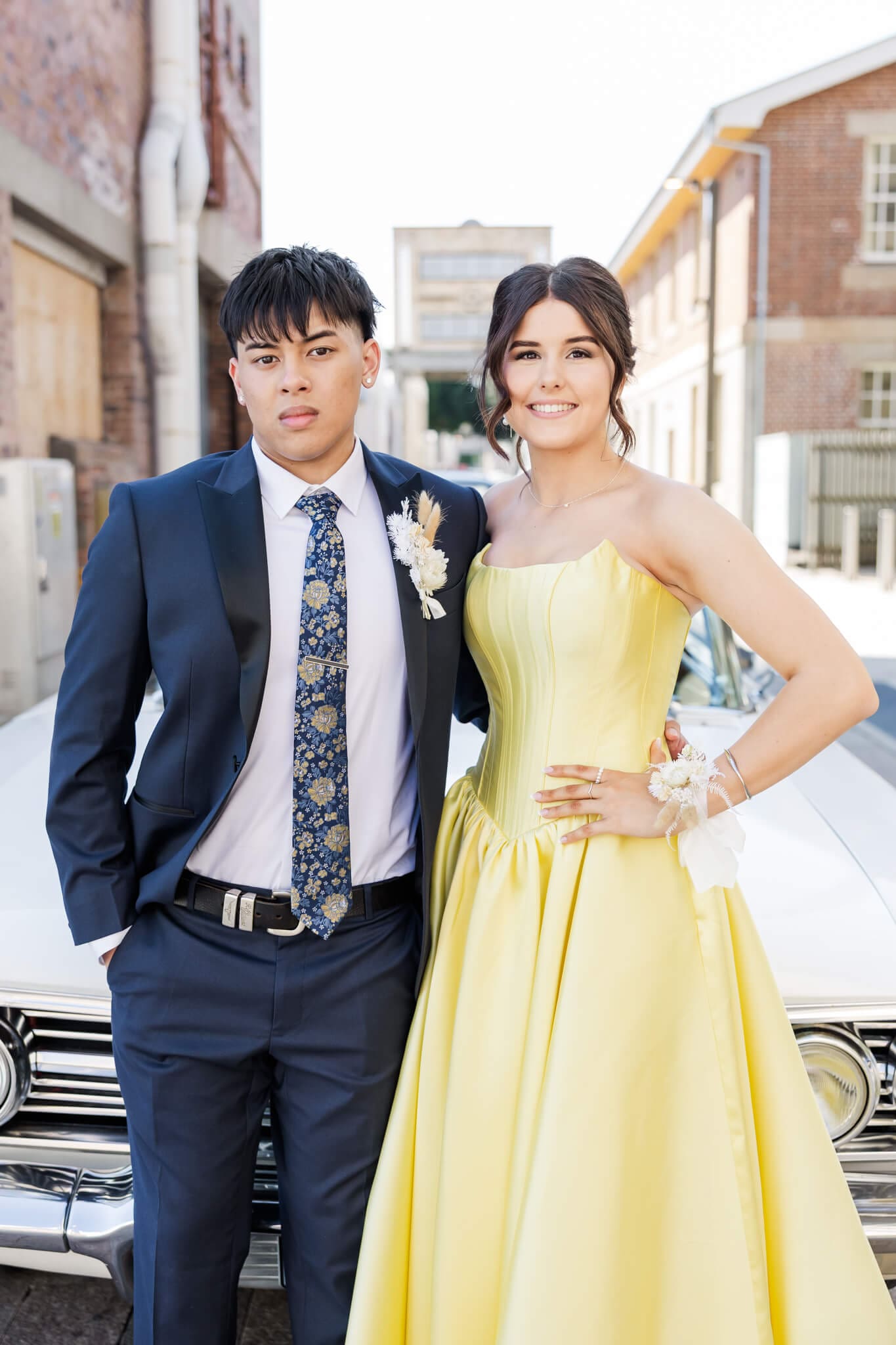 A couple of graduates, a boy and girl, posing for their formal photo in Rockhampton in front of a white old fashioned car.