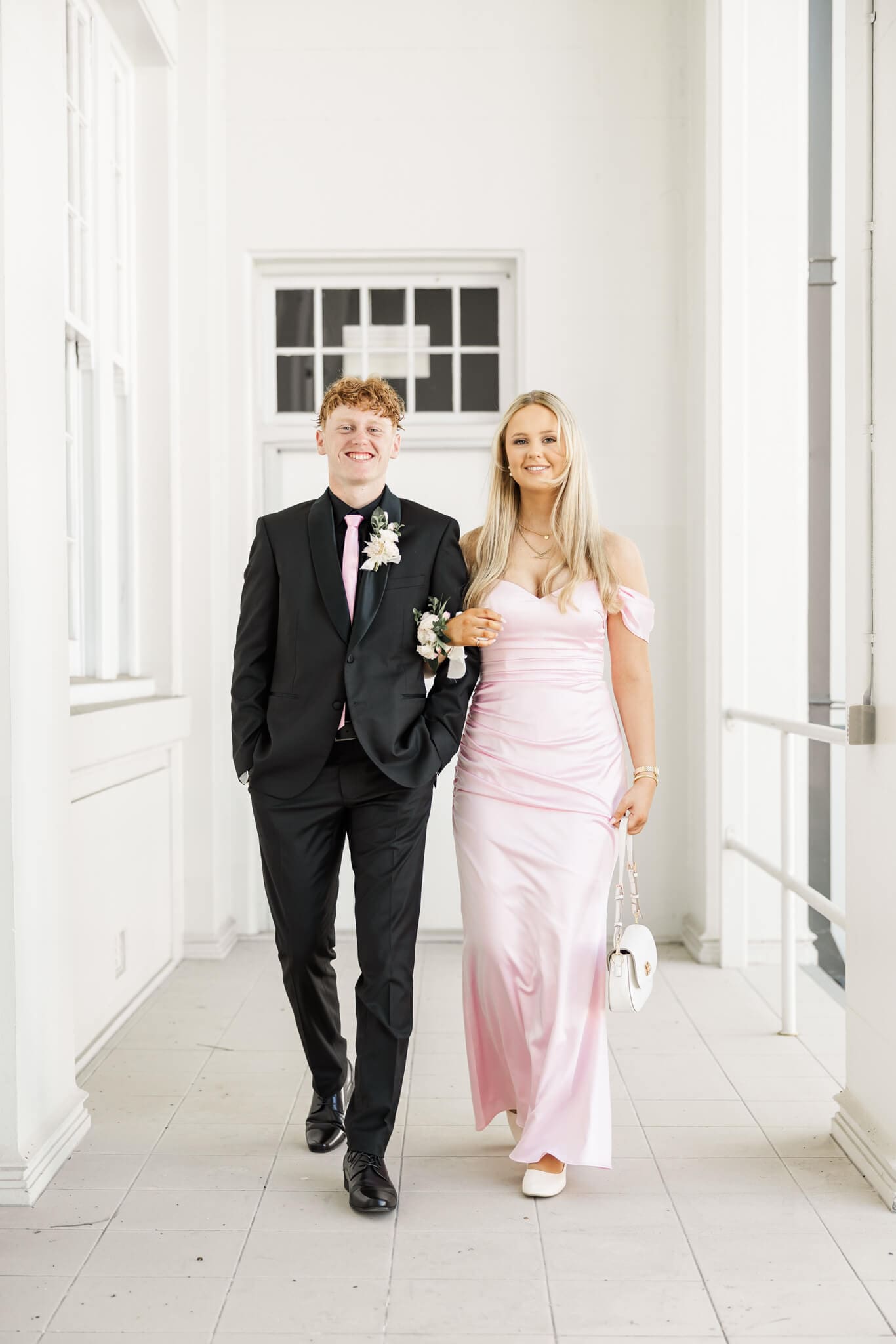 Formal photos of two graduates as a couple, walking towards the camera. Girl is wearing a beautiful pink dress and guy is in a black suit.