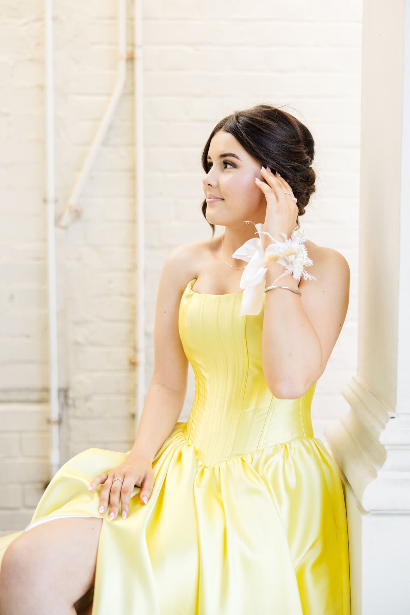 A young girl in a beautiful yellow dress wearing a hand wrist floral piece created by Shannon Hawkes Rockhampton, sitting on a balcony wall, posing for her formal portraits.