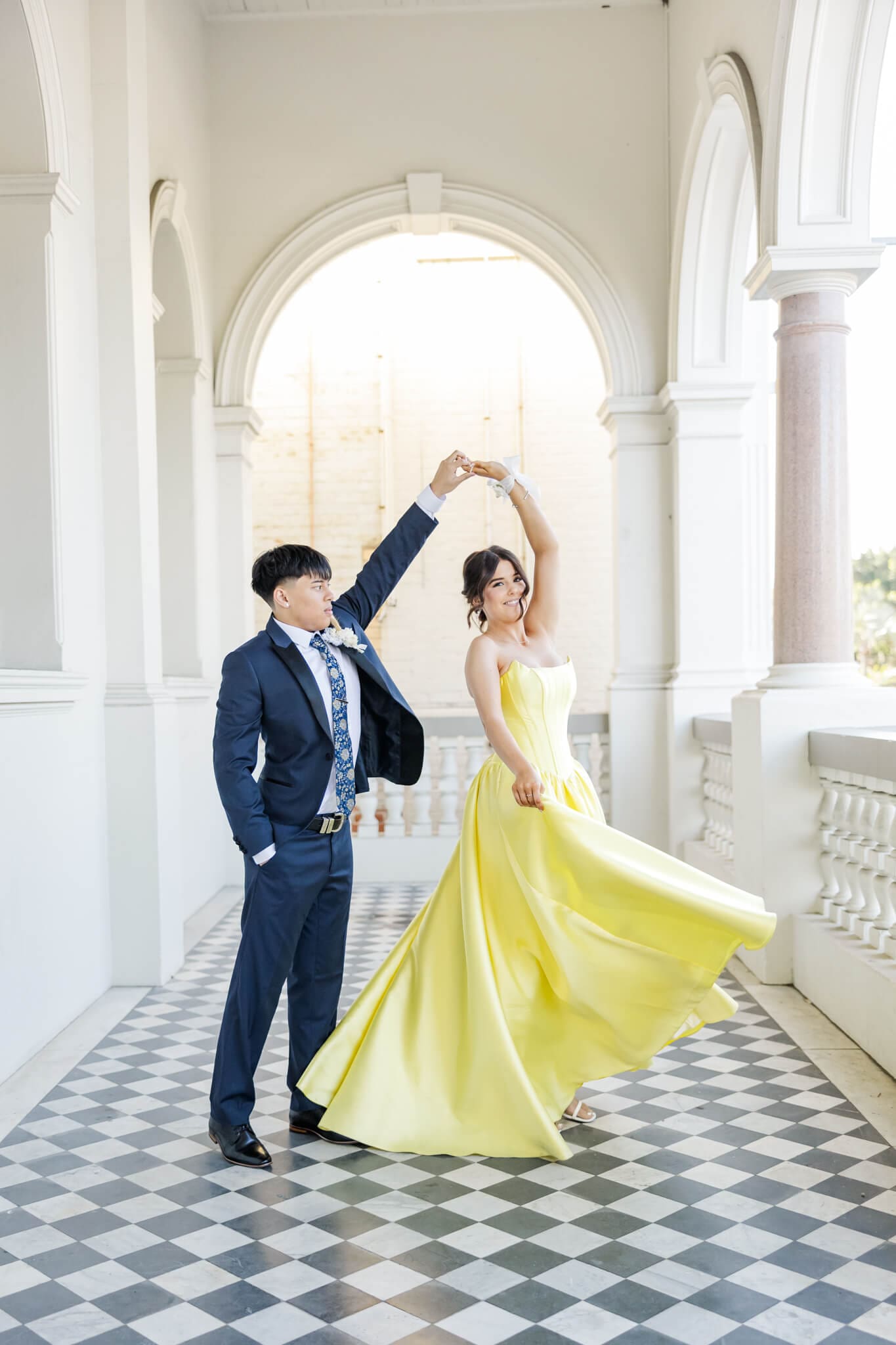 Boy graduate twirling girl graduate in a gorgeous old building with a checkered black floor. Boy is dressed in a navy suit and the girl is dressed in a beautiful yellow dress which flares out as she twirls.
