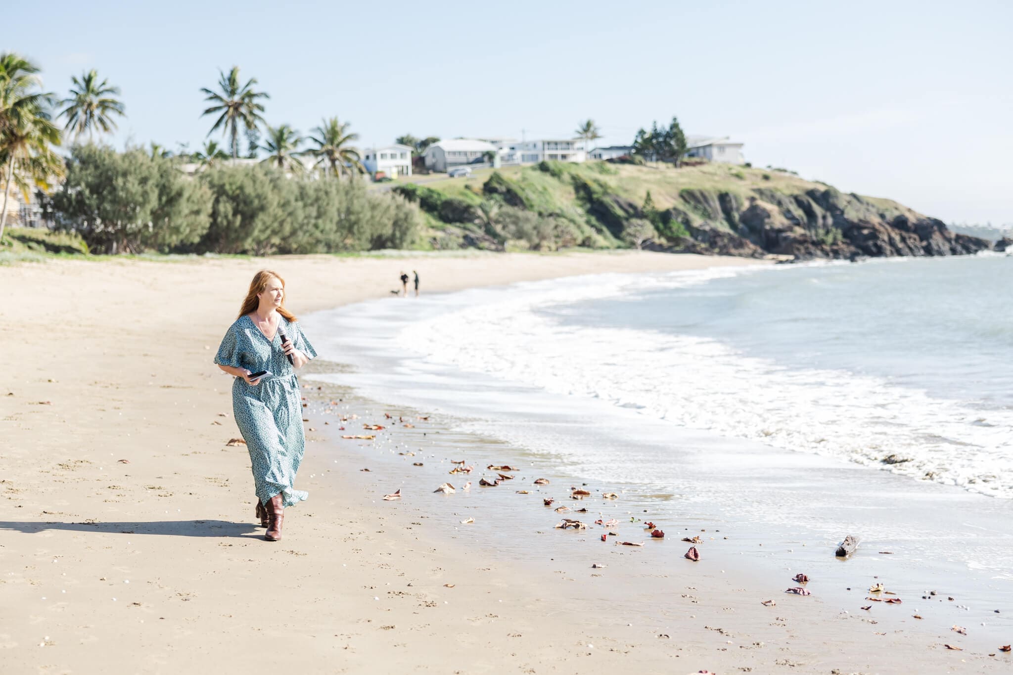Marriage Celebrant walking along a Yeppoon beach in a green dress holding a microphone and a notebook.