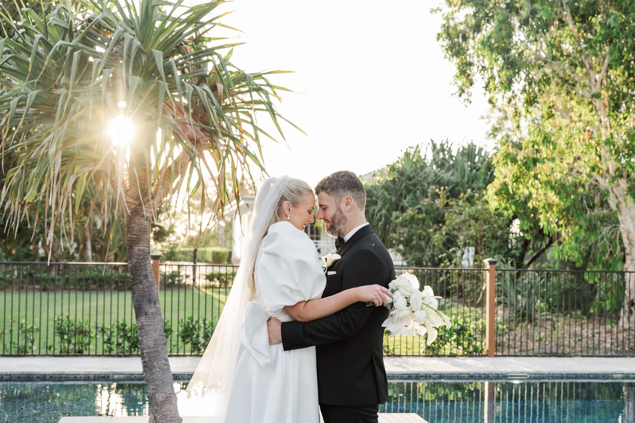 Gorgeous photo of the bride and groom with the sun glowing behind them while they stand next to a beautiful pool at a luxury venue.