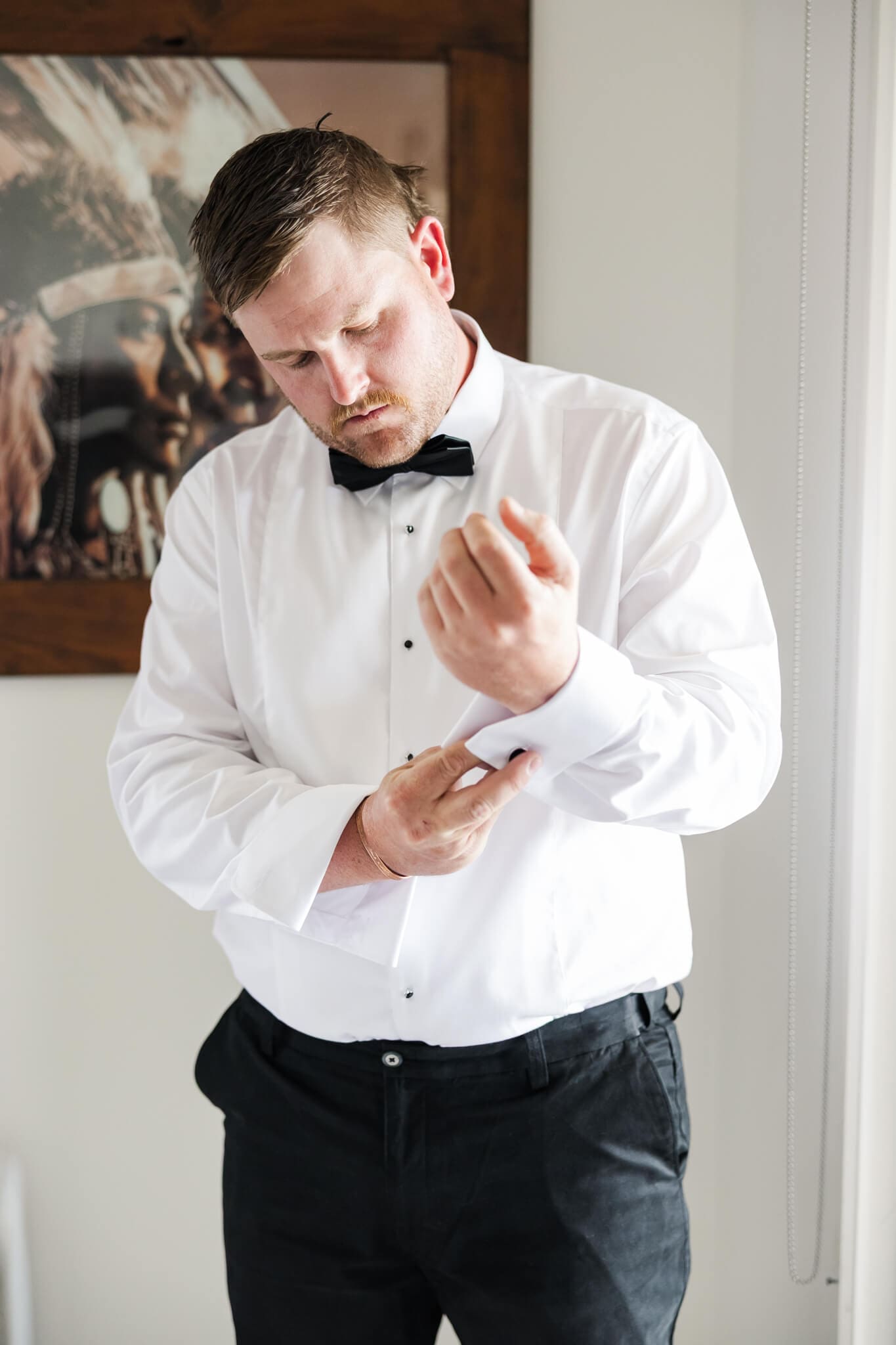 A groom looks down as he adjusts a cuff link on his long sleeve shirt.