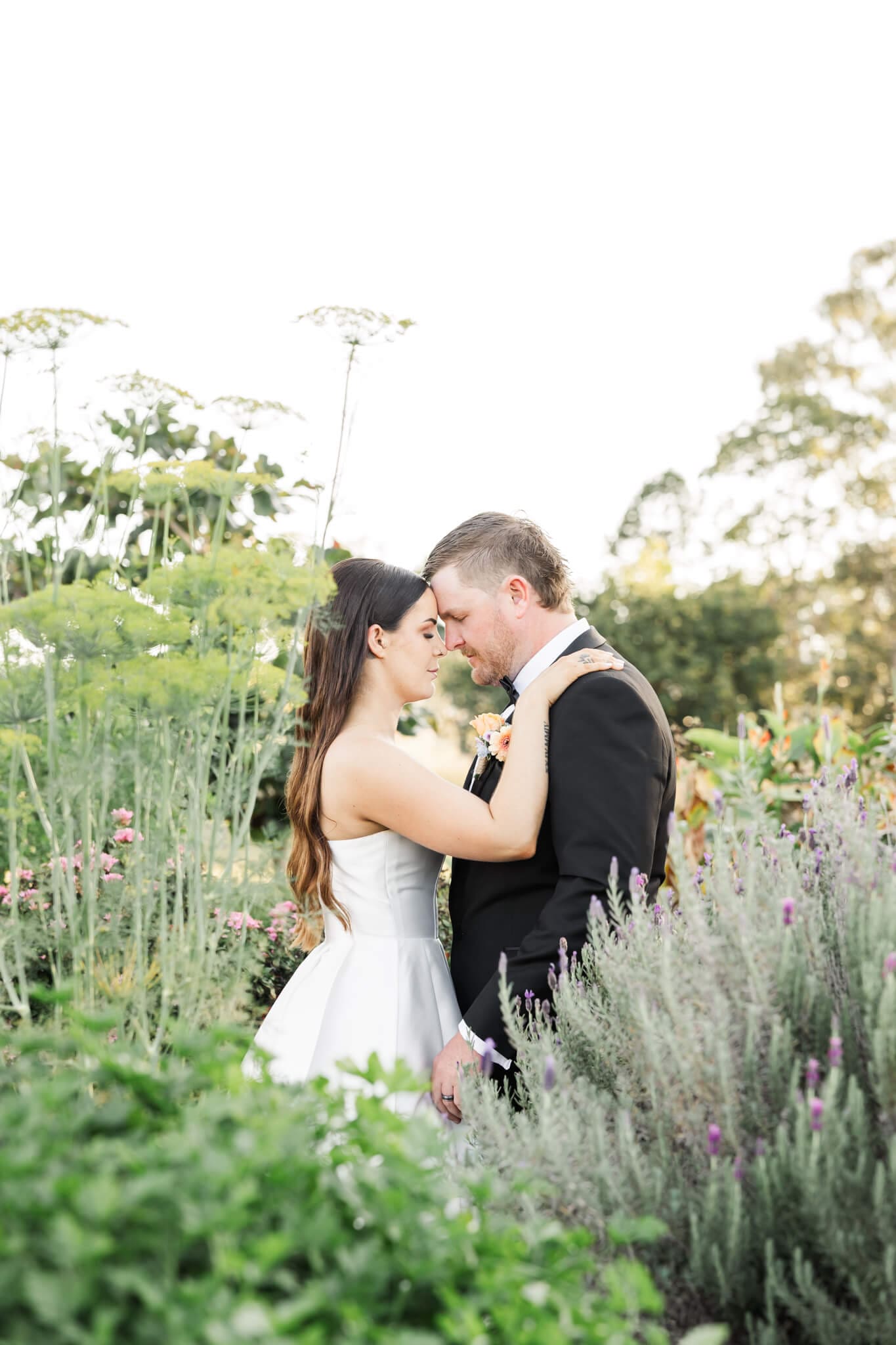 Bride and Groom stand amongst beautiful gardens, their heads touching. Captured by Yeppoon Photographer Julie-Anne Photography.