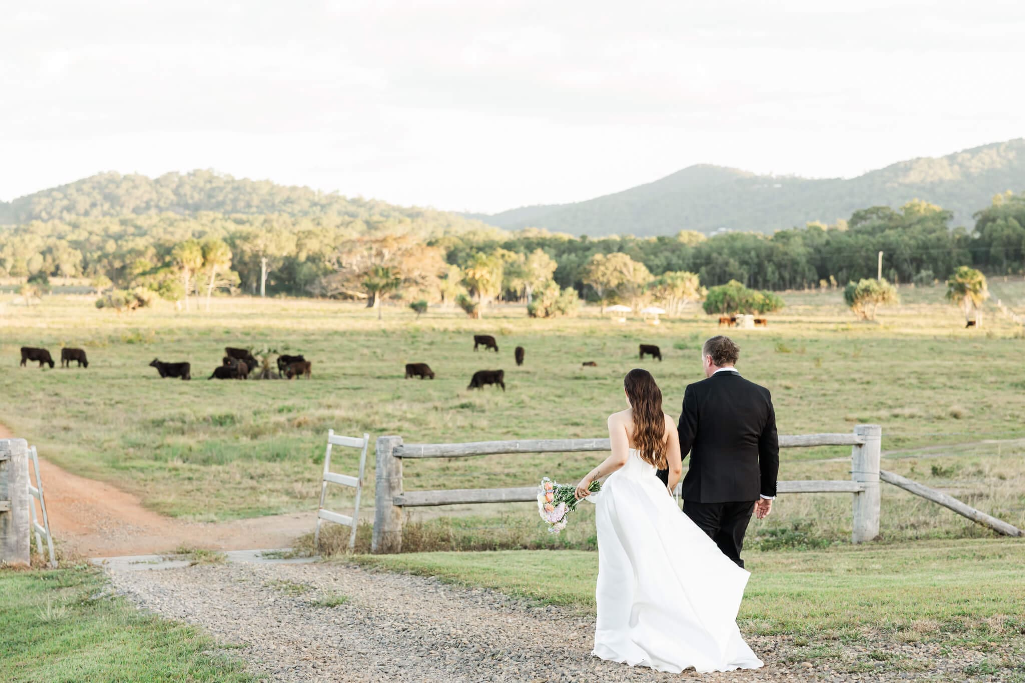 Bride and groom walking down a gorgeous countryside dirt road towards an open paddock at sunset.