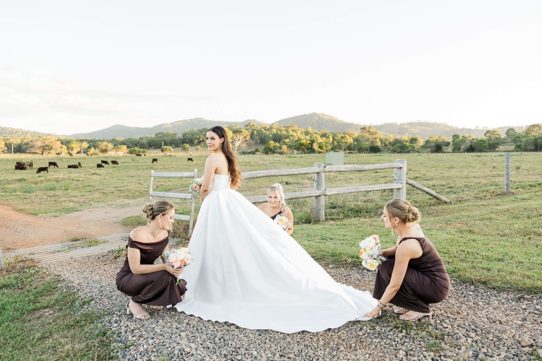 A bride allows her bridesmaids fix her dress and long train for a photo on a gravel road in the countryside.