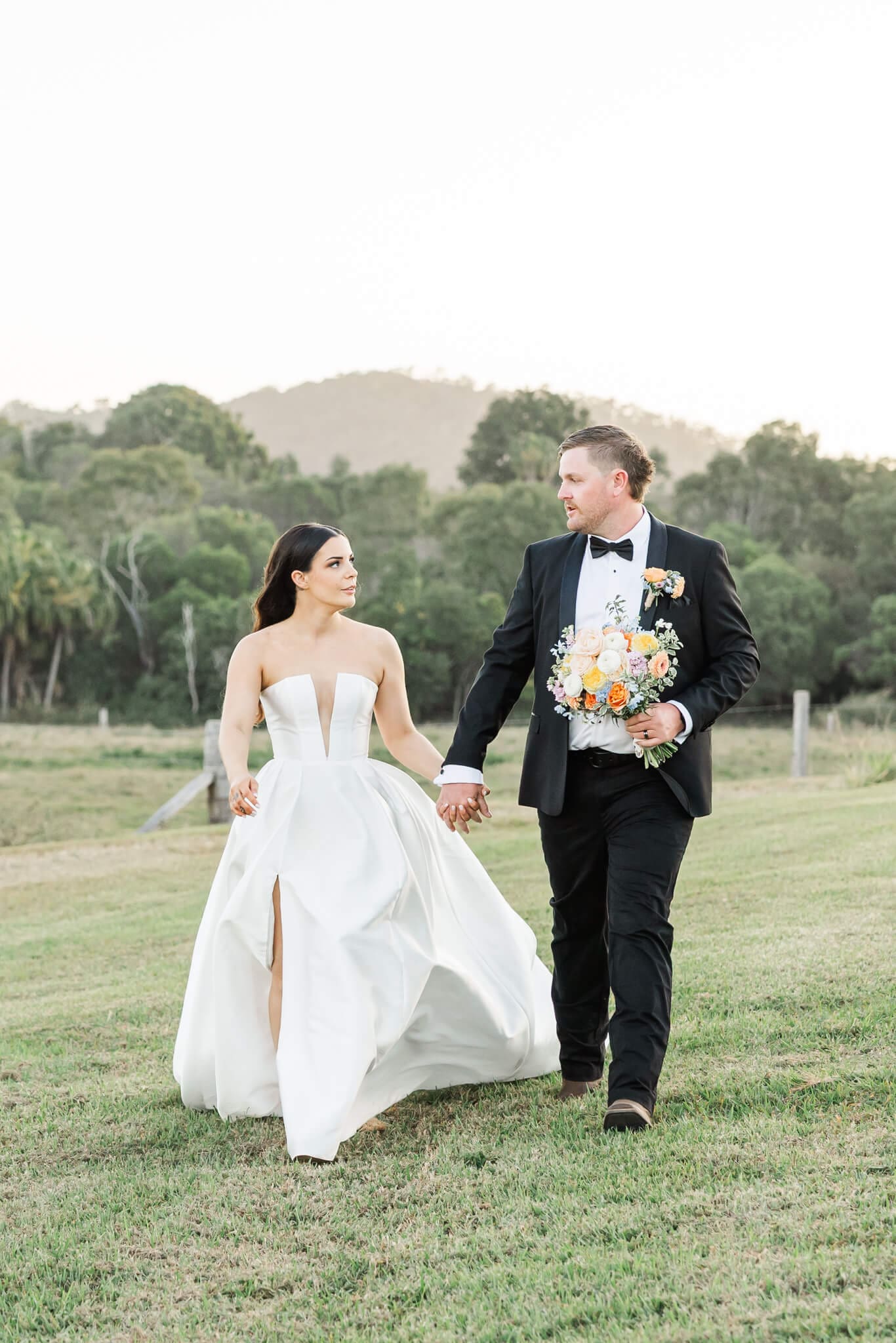 The groom carries the bouquet and holds the brides hand with his other hand as they walk freely across an open paddock.