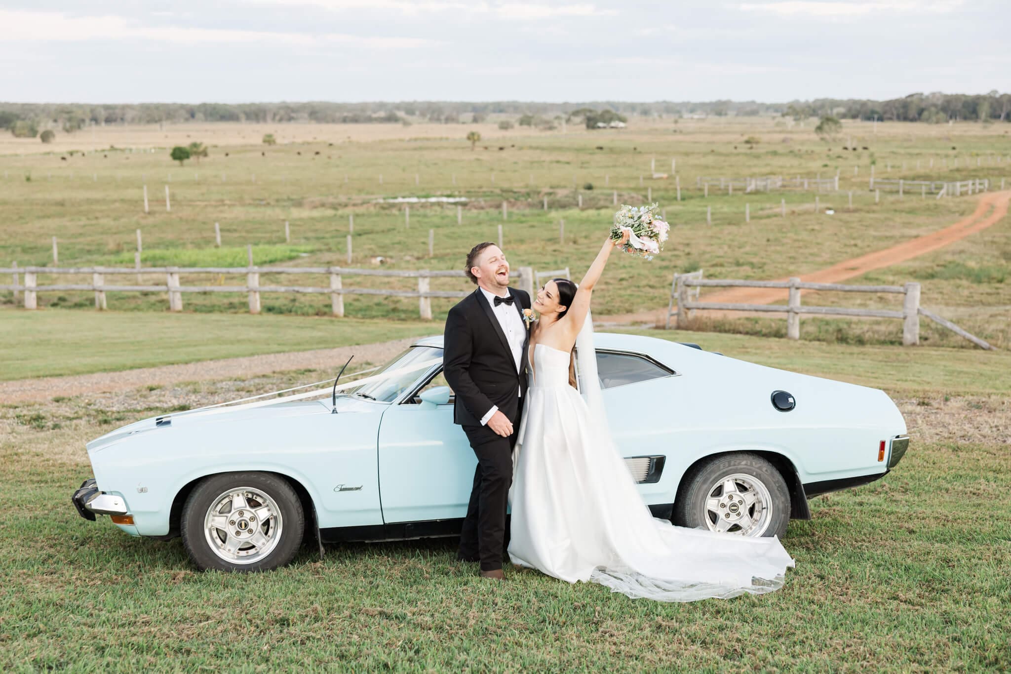 Bride and Groom standing next to an old blue car in an open paddock, the bride throwing her bouquet in the air cheering while the groom looks on amused.