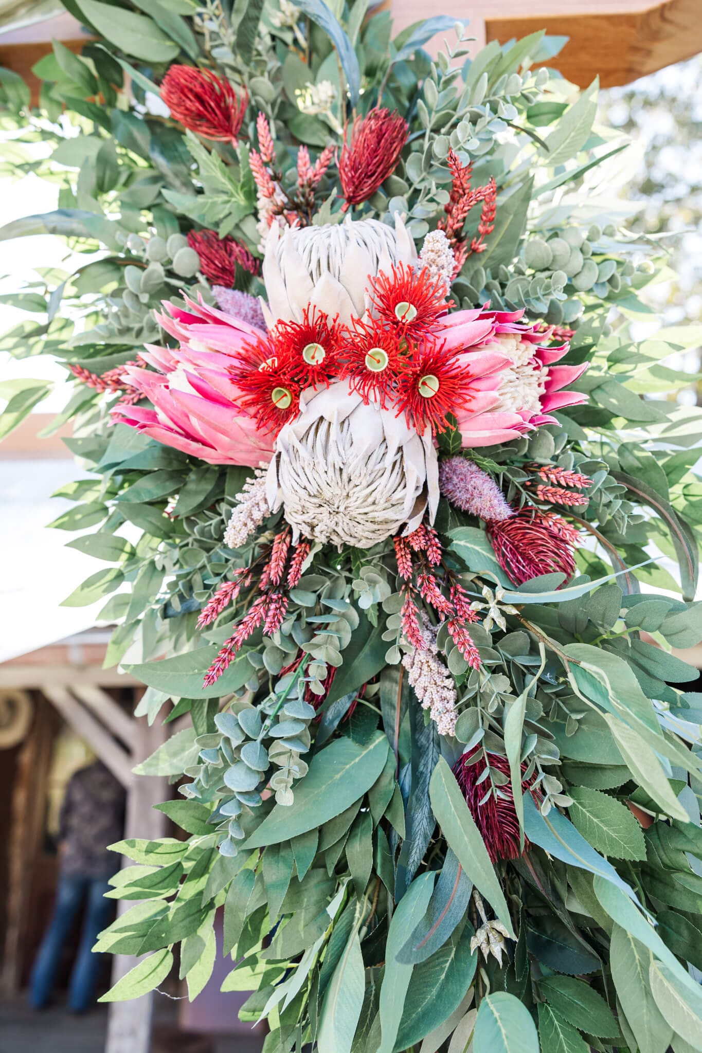 A beautiful floral piece hanging on a wedding arbor, featuring Australian native flowers that are pink and red, surrounded by green native leaves.