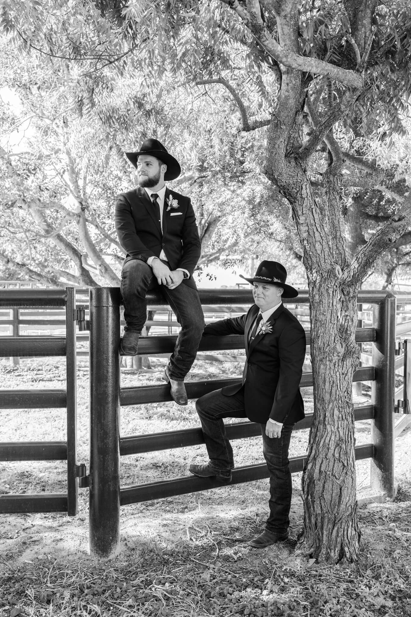 Black and white image of a groom sitting on the top rail of a set of yards while his best man stands below, both looking off into the distance.