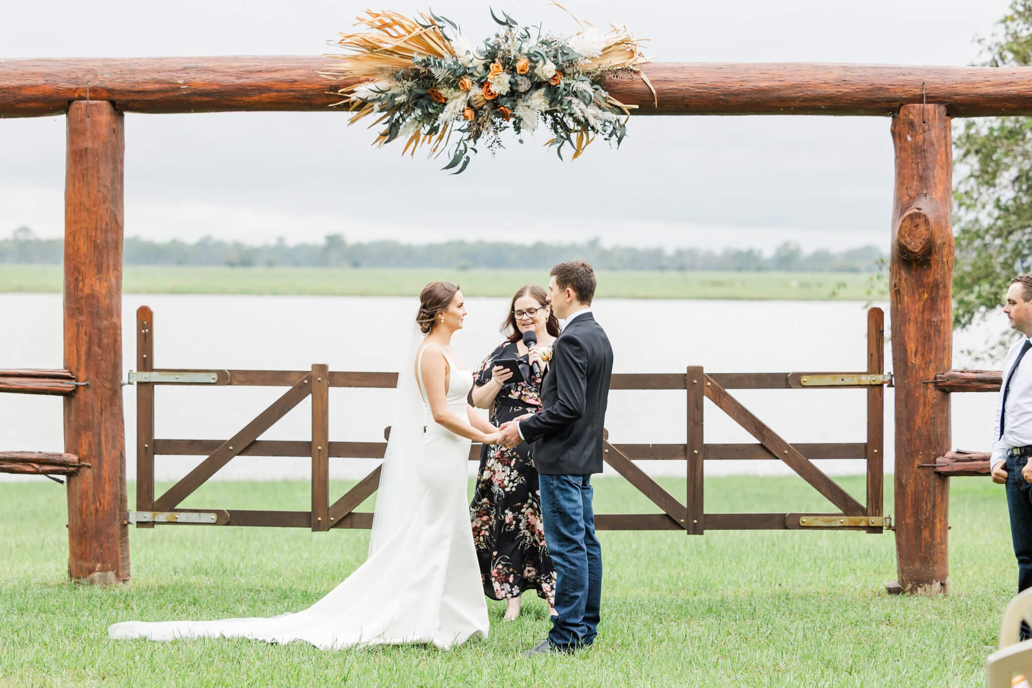 Wedding ceremony at Paradise Lagoons Rockhampton. The bride and groom exchange vows in front of a set of rails and gates next to a lagoon.
