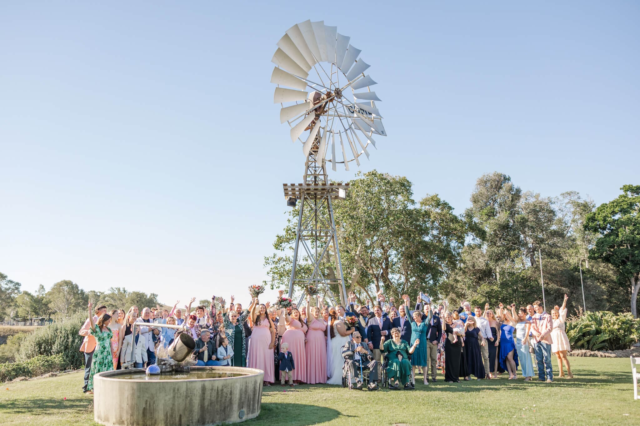 Friends and families celebrate as the bride and groom kiss. There is a windmill in the background and an Australian landscape.