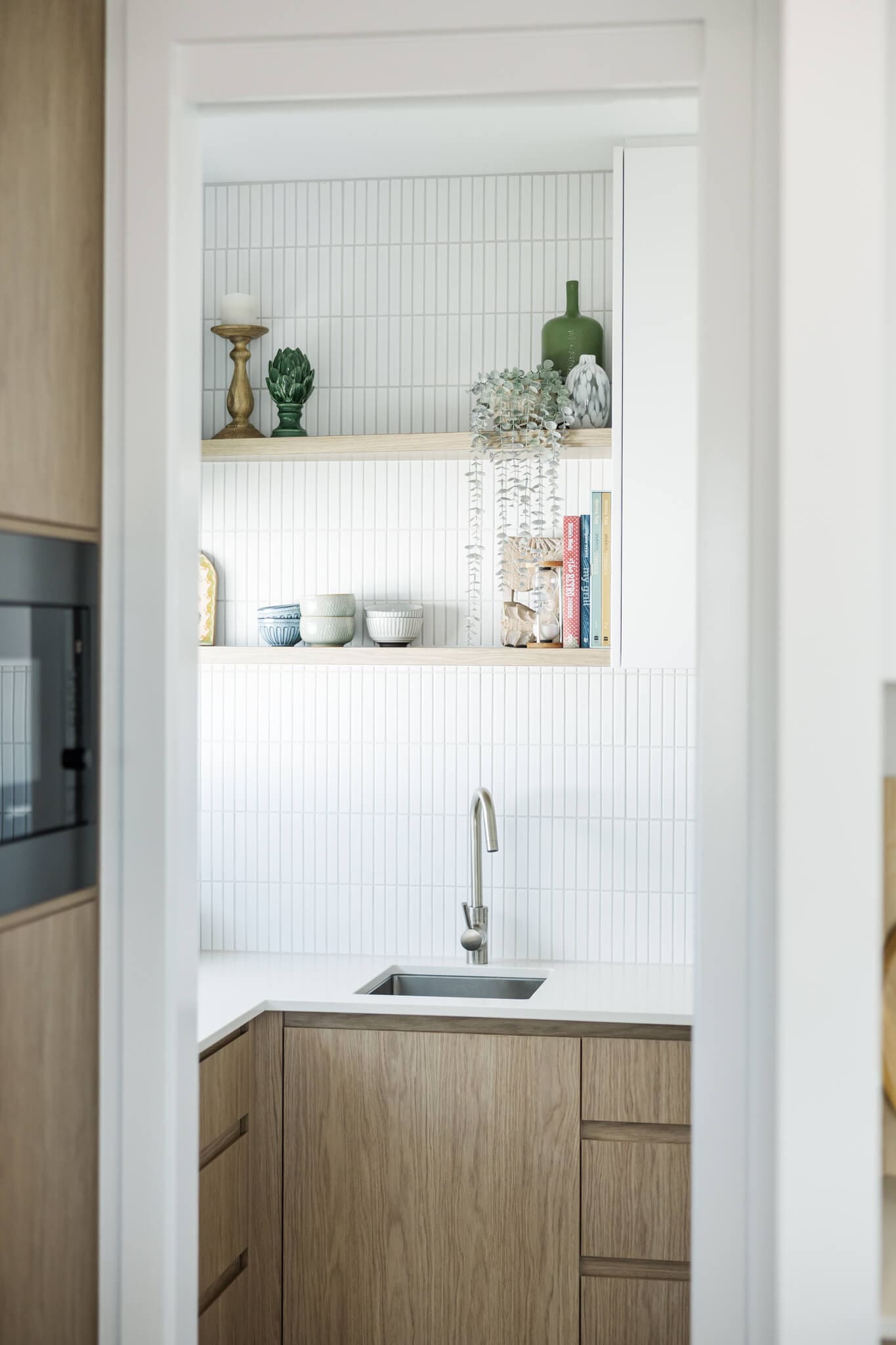 Beautiful details in an Airbnb pantry area including cook books, vases and a plant falling over the shelves which are a light coloured wood against white tiles.
