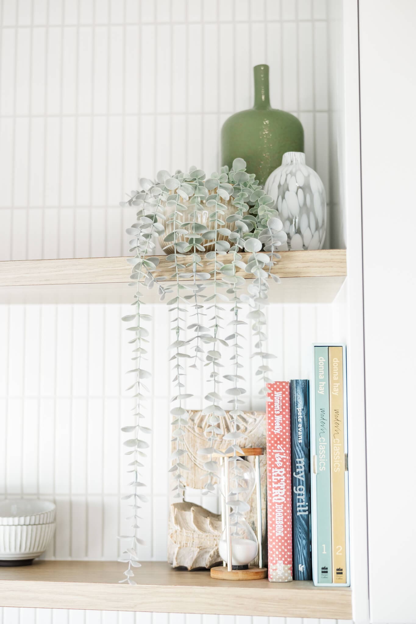 Wooden shelves with cook books, vases and a plant falling over the shelves against white tiles.