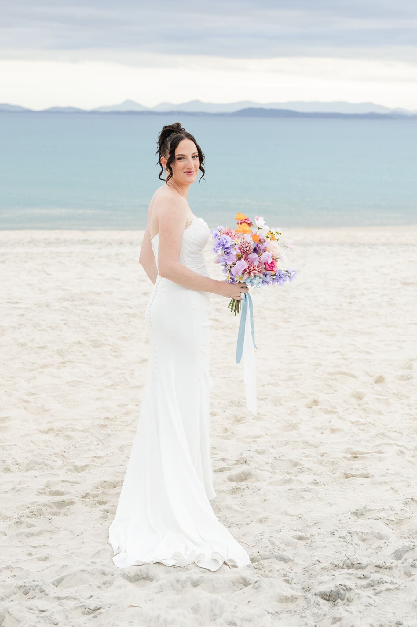 Beautiful bride looks back over her shoulder as she stands on a beautiful beach. She stands in a stunning white dress, which has had dress alterations in Rockhampton.
