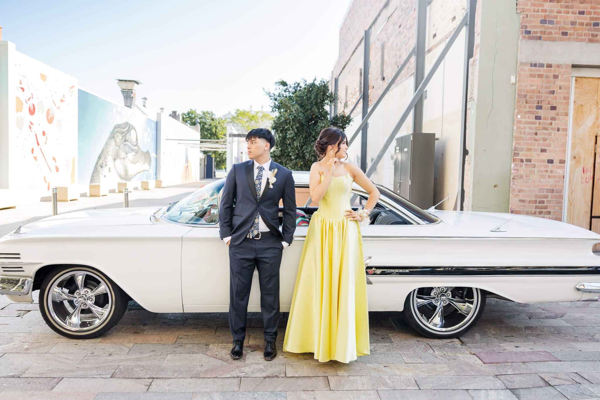 Formal graduates, a boy and a girl, stand in front of their hire care for their photography session. She wears a stunning yellow gown.