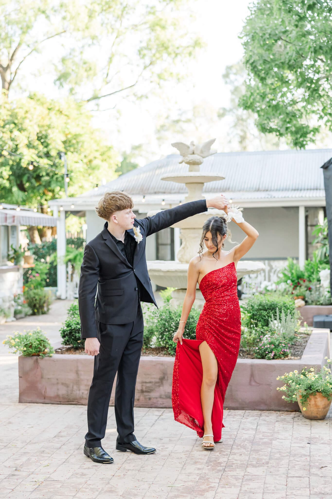 Graduate boy spins girl for their formal photos. Girl wears a stunning red slim dress and he is dressed in a black suit. Captured by Julie-Anne Photography.
