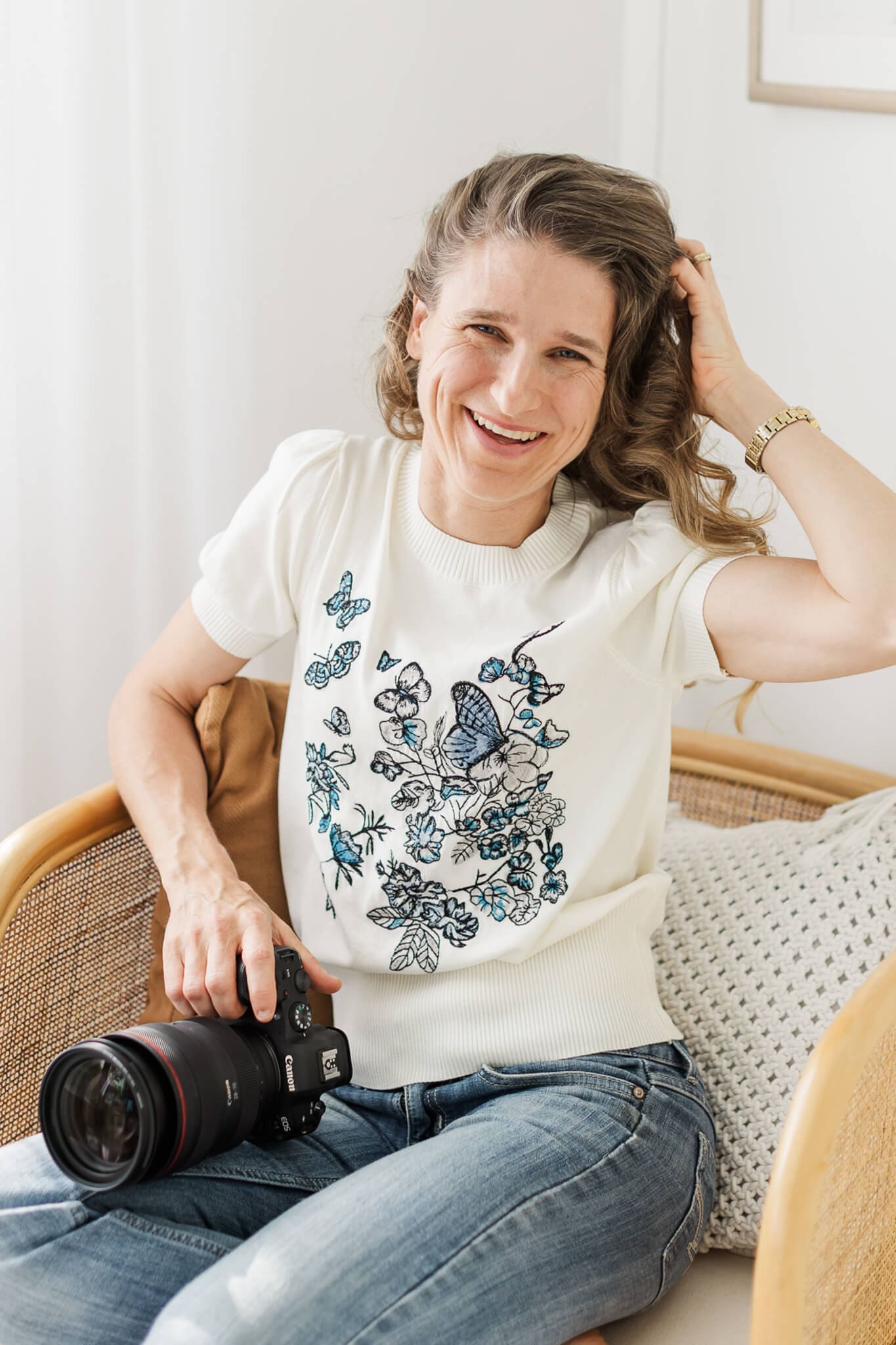 Branding photo of Julie-Anne Photography. She is holding her camera and sitting in a cane chair, smiling happily at the camera.