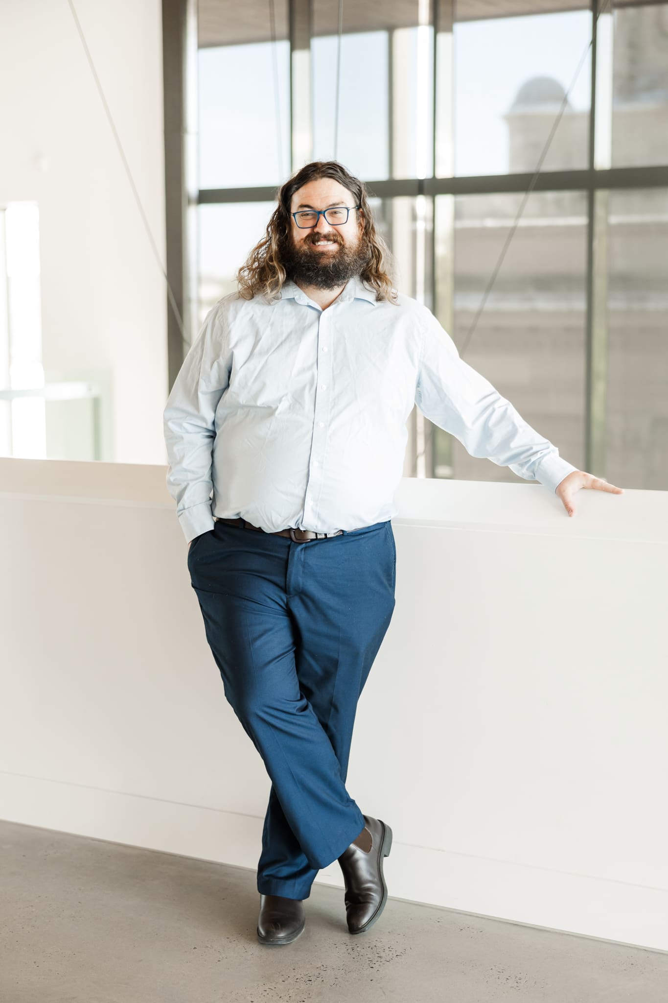 A man stands leaning against a cream balcony wall, wearing a blue shirt with one leg crossed over the other.