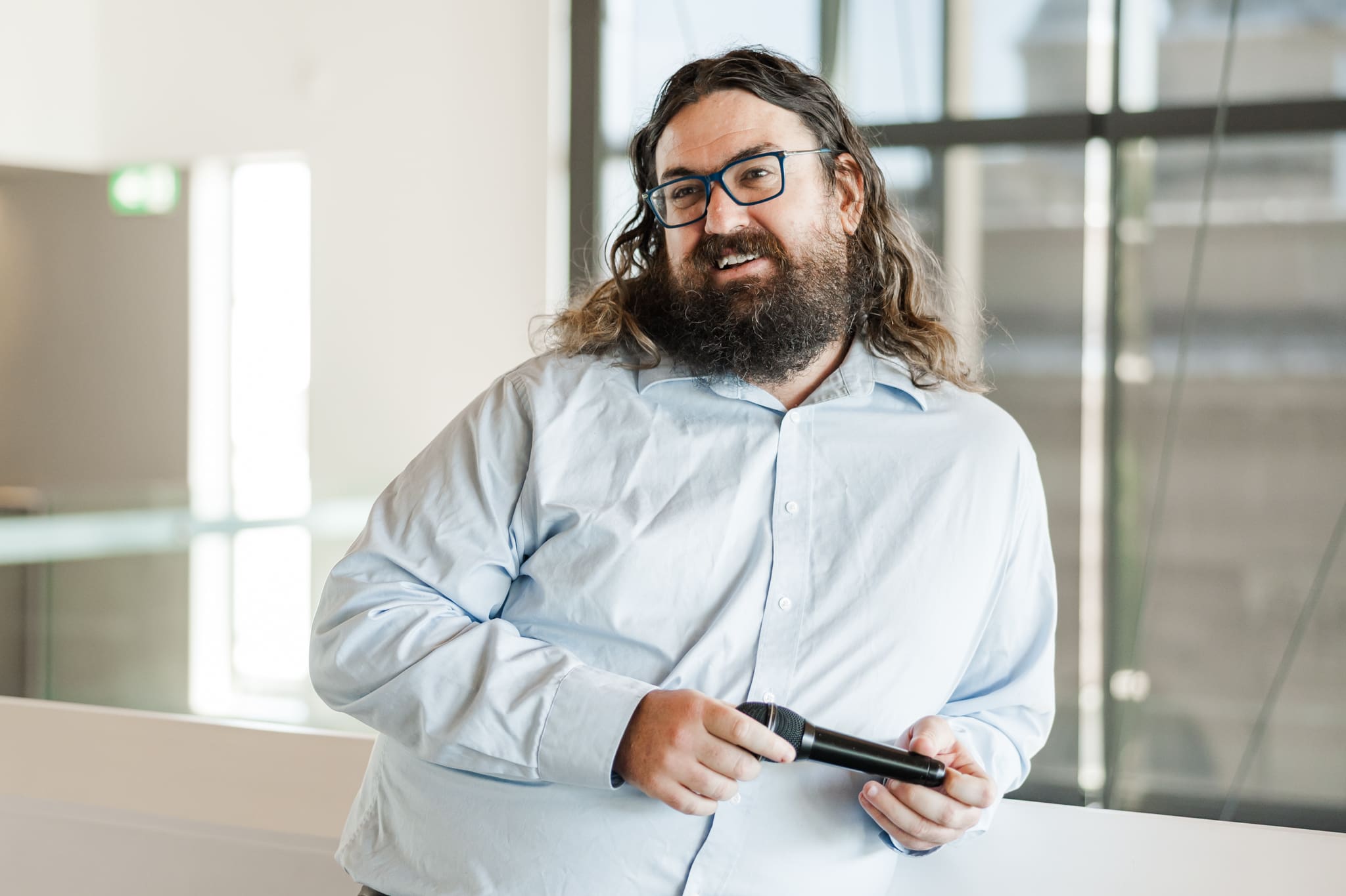A man stands with a mic in his hands, leaning against a cream balcony wall, wearing a blue shirt.