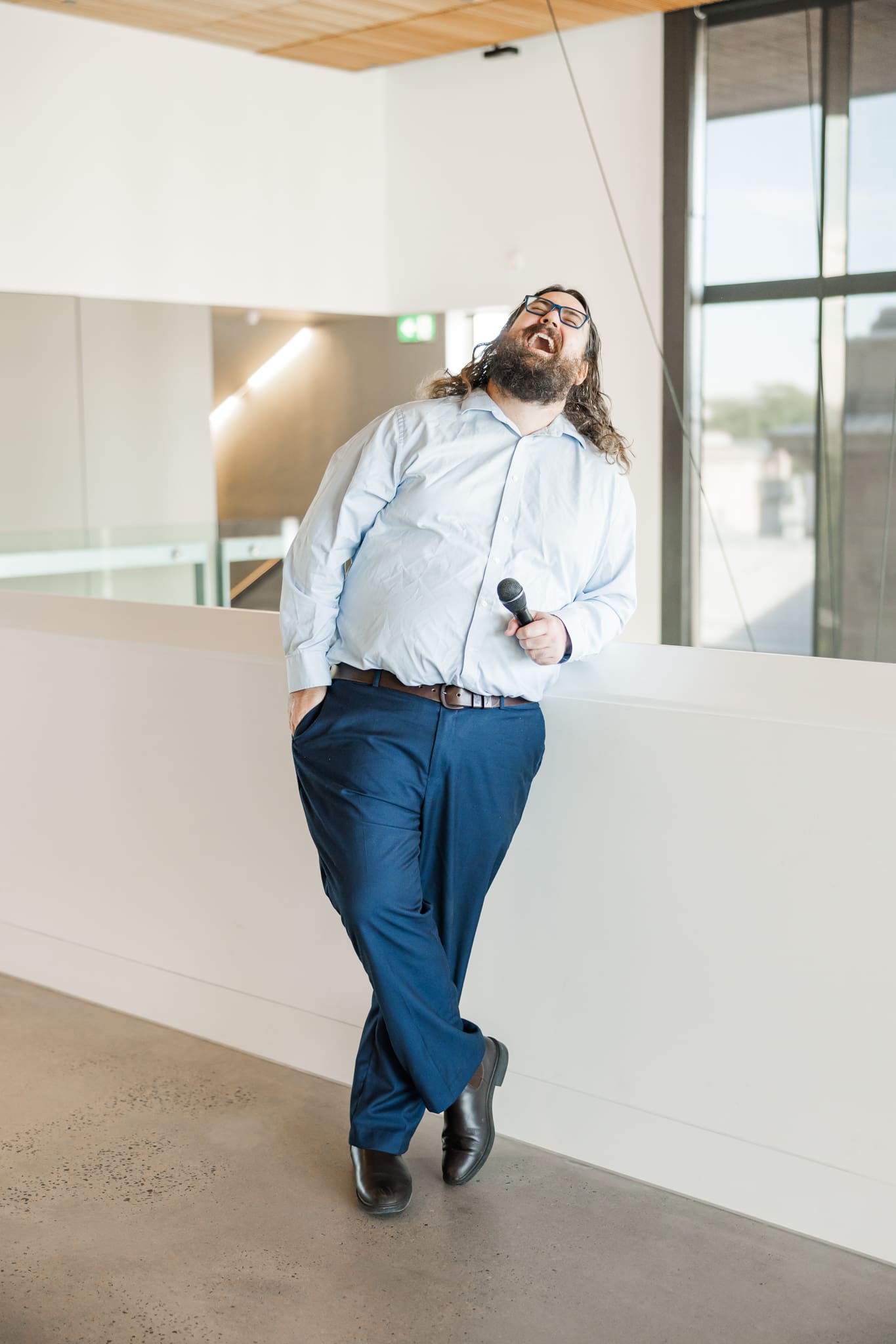Married by Mitchell, a marriage celebrant, laughs towards the ceiling as he stands near a cream wall with a mic in his hands.