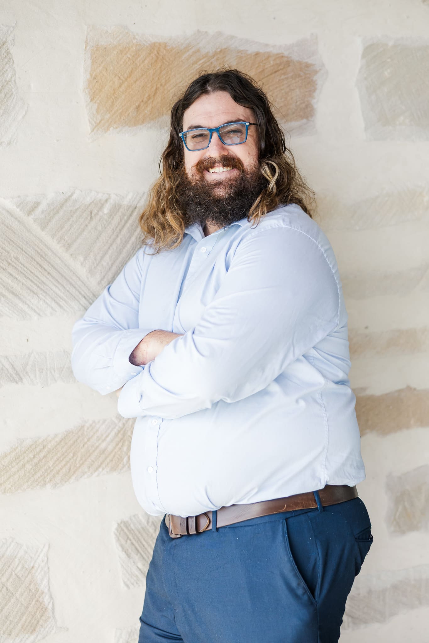 A man leans against a cream brick wall, looking towards the camera, dressed in a blue shirt and blue pants.