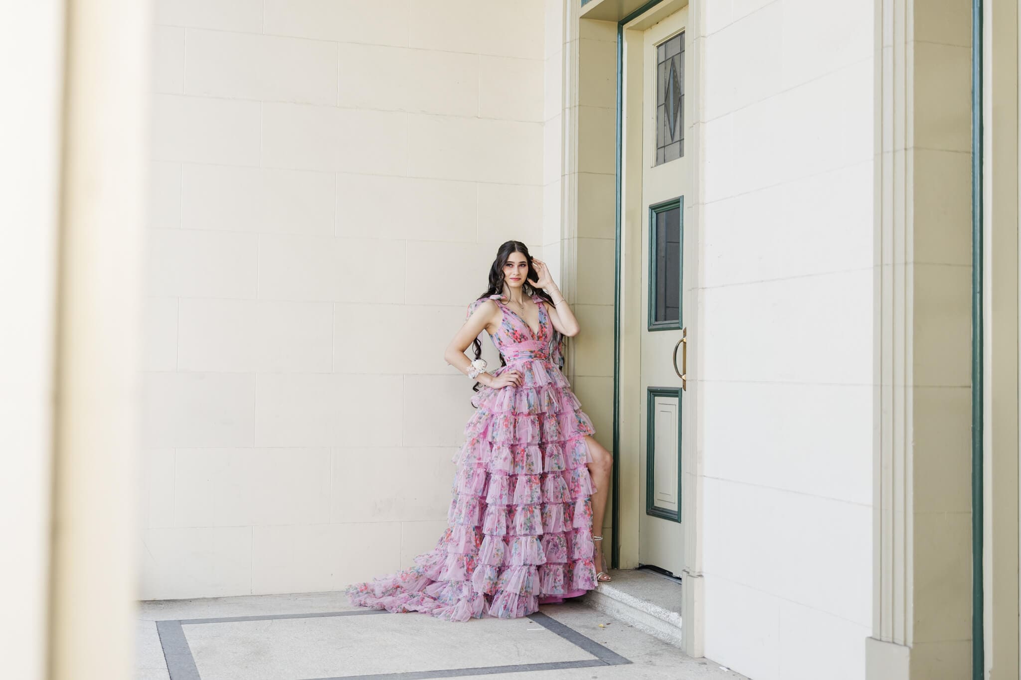 High school graduate poses in a cream building wearing a stunning [ink, frilly, Oak and Valentine gown.