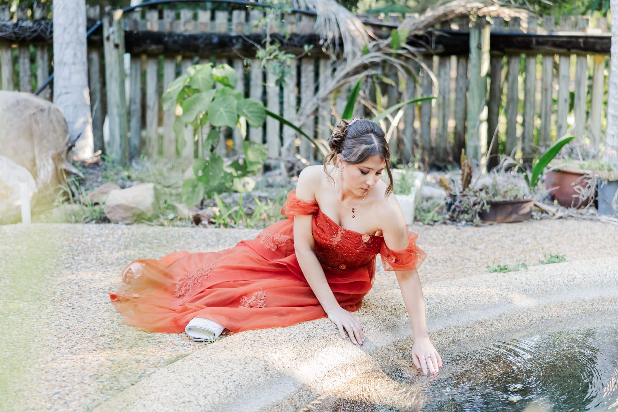Grade 12 girl poses for her school formal photos, seated beside a pond running her fingers through the water.