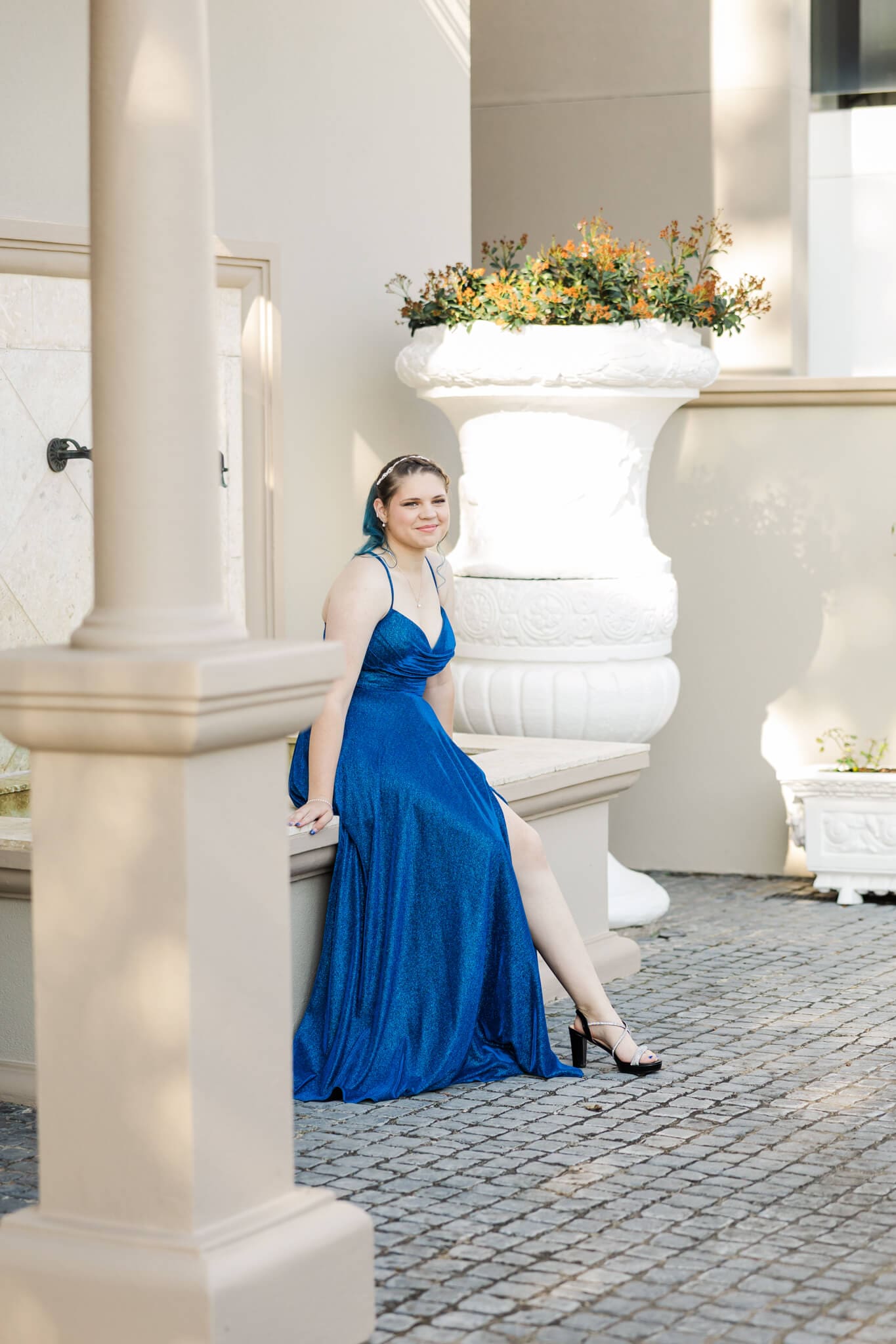 Beautiful portrait of a high school senior girl. She sits in a courtyard in a royal blue dress with black high heels on.