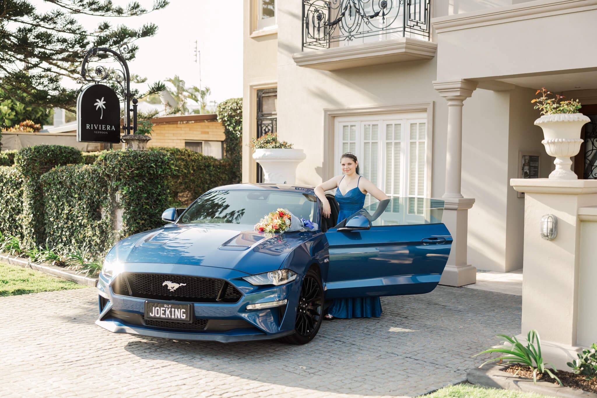 Young girl poses for her school formal photos while she stands next to a blue mustang with one hand on her hip and a bunch of flowers on the bonnet of the car.