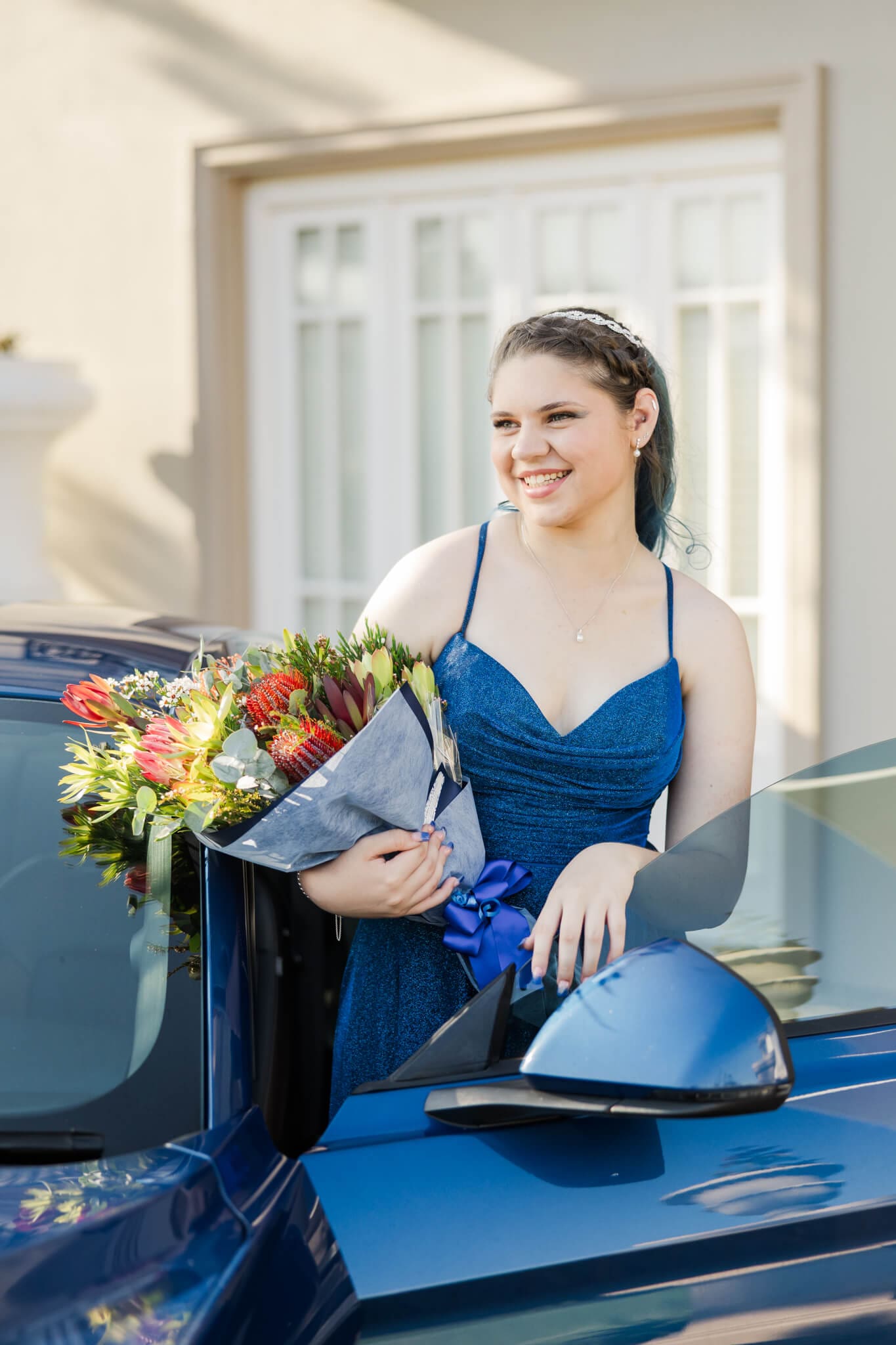 Young girl poses for her school formal photos while she stands next to a blue mustang with a bunch of flowers in her hands, smiling off to the distance.