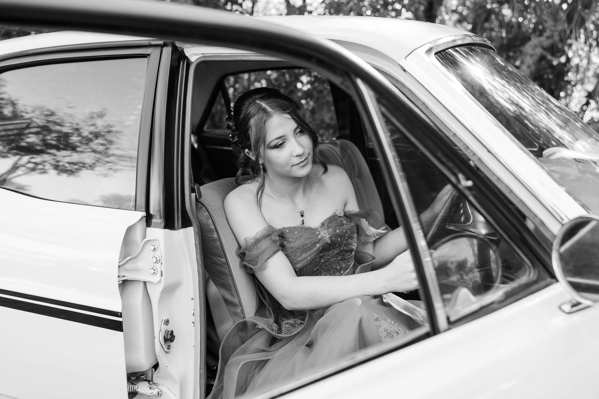 Black and white image of a girl looking out the front of an old fashioned sedan car for her school formal photos.