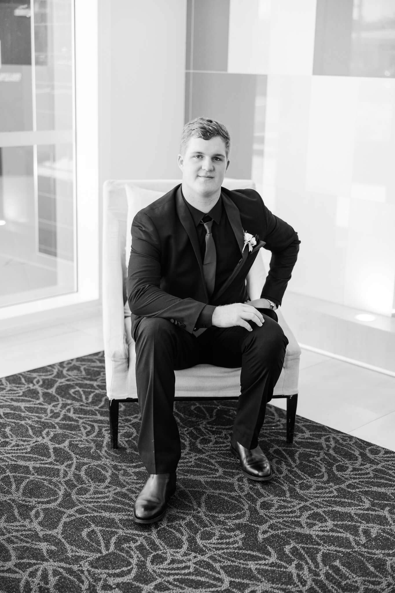 Black and white image of high school senior boy sitting on a chair, elbow on knee, looking towards camera.