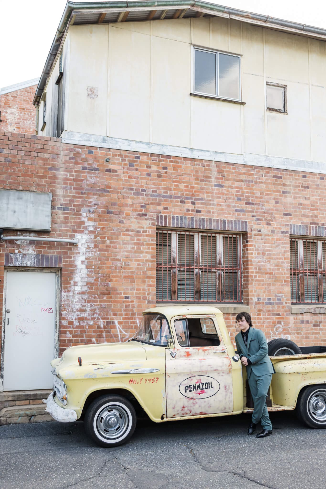 High school senior stands near an old holden yellow ute which is parked in an alleyway next to an orange bricked building.